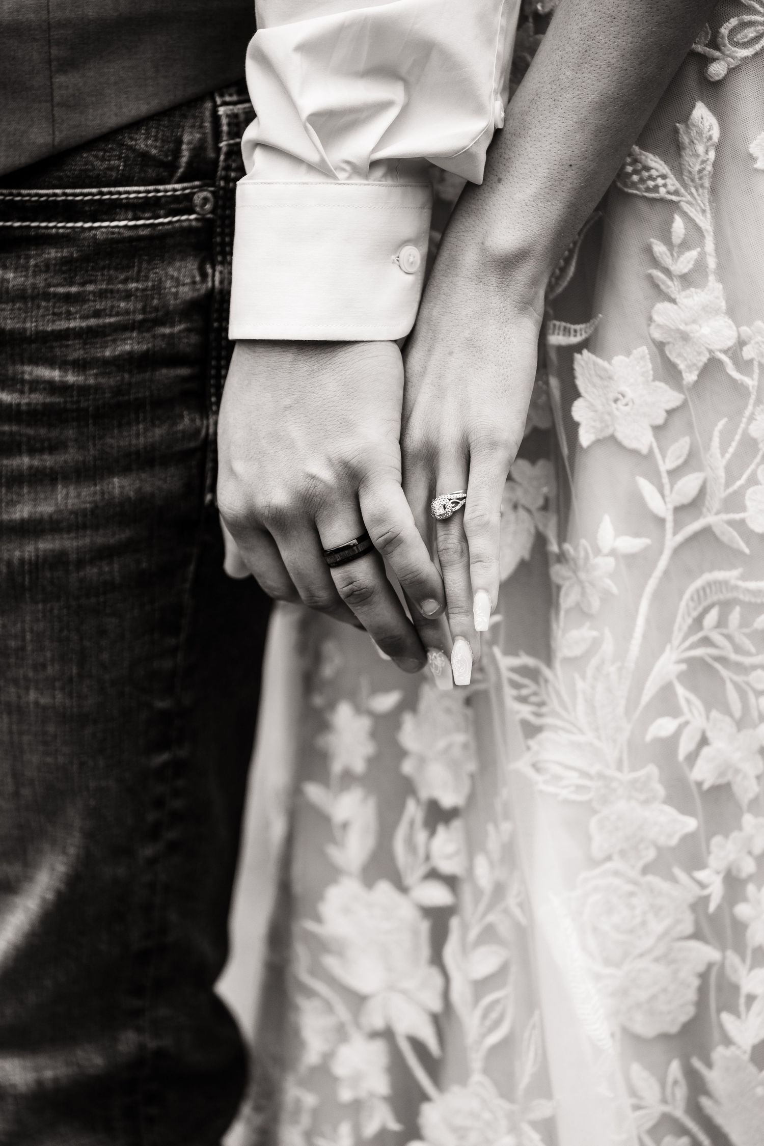 Black and white close-up of couple's hands with engagement ring against a lace dress.