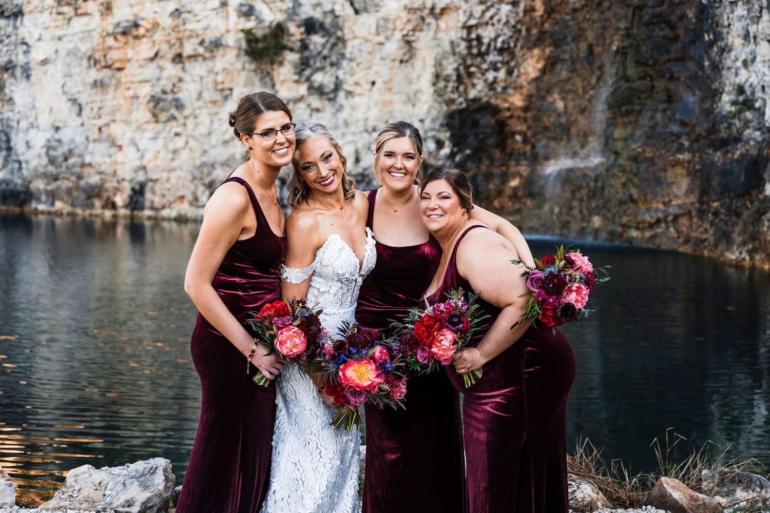Wedding party in burgundy dresses posing by a scenic waterfront.