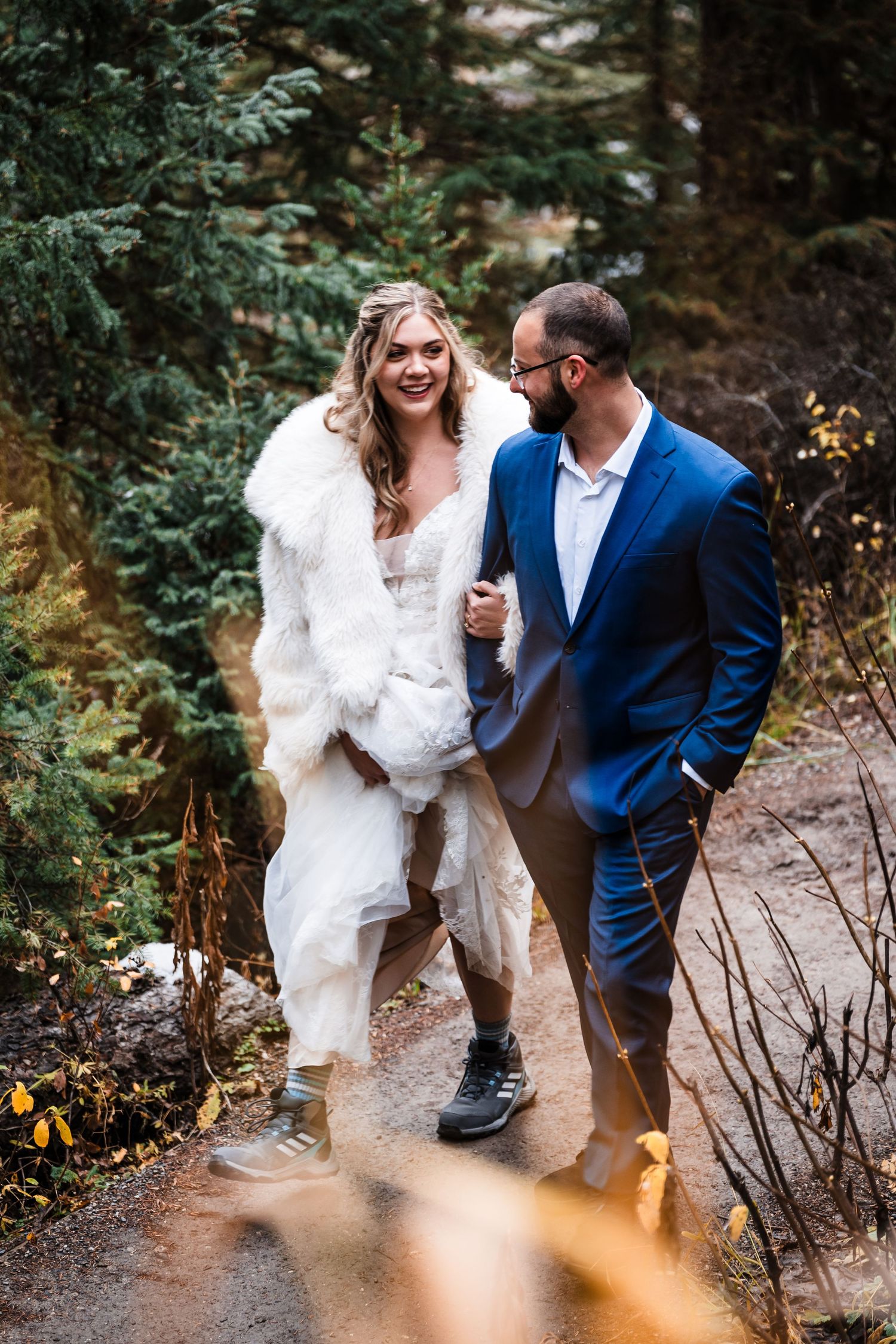 Couple in wedding attire walking through an evergreen forest path.