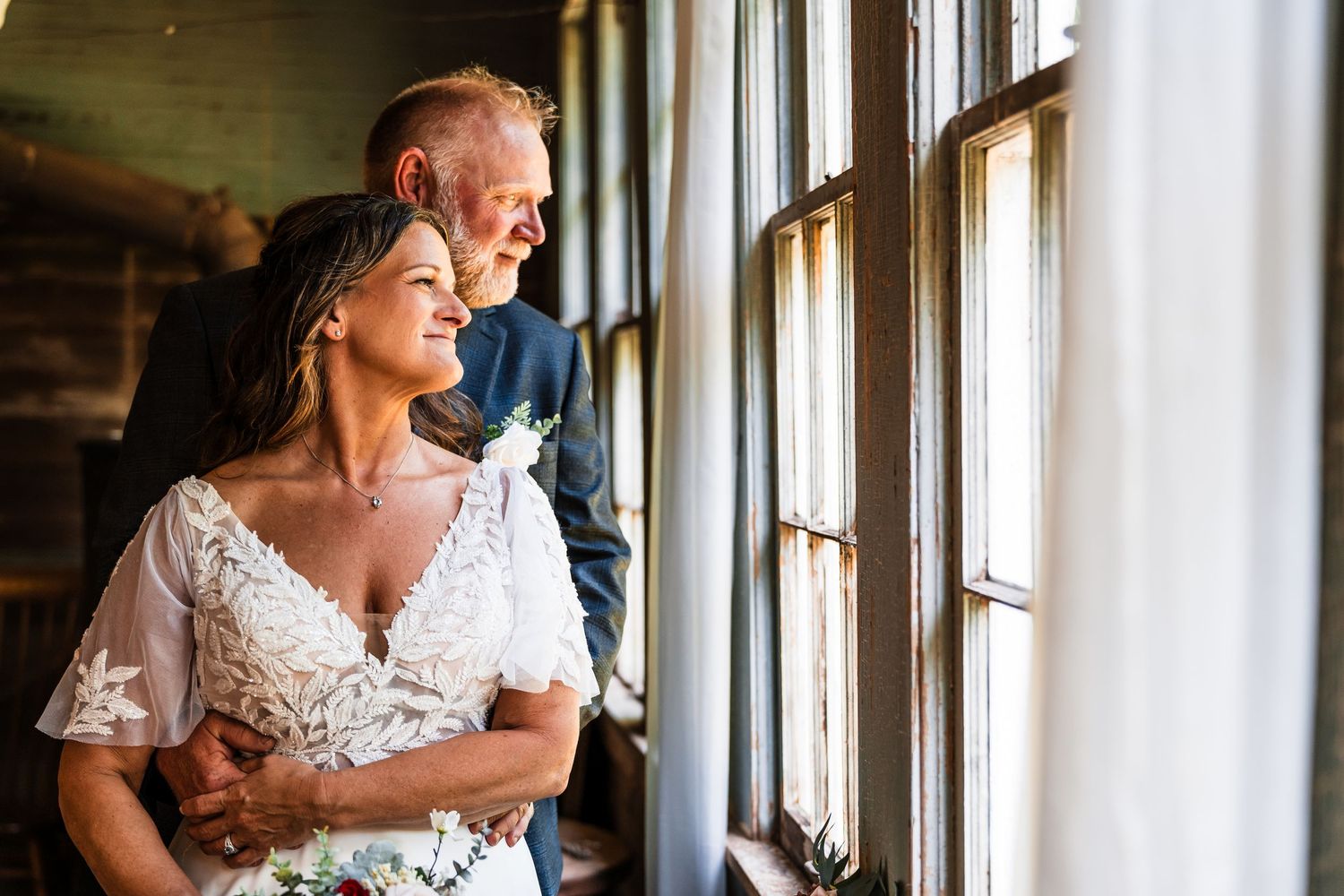 A tender wedding moment is shared in front of sunlit windows with soft natural light illuminating the scene.