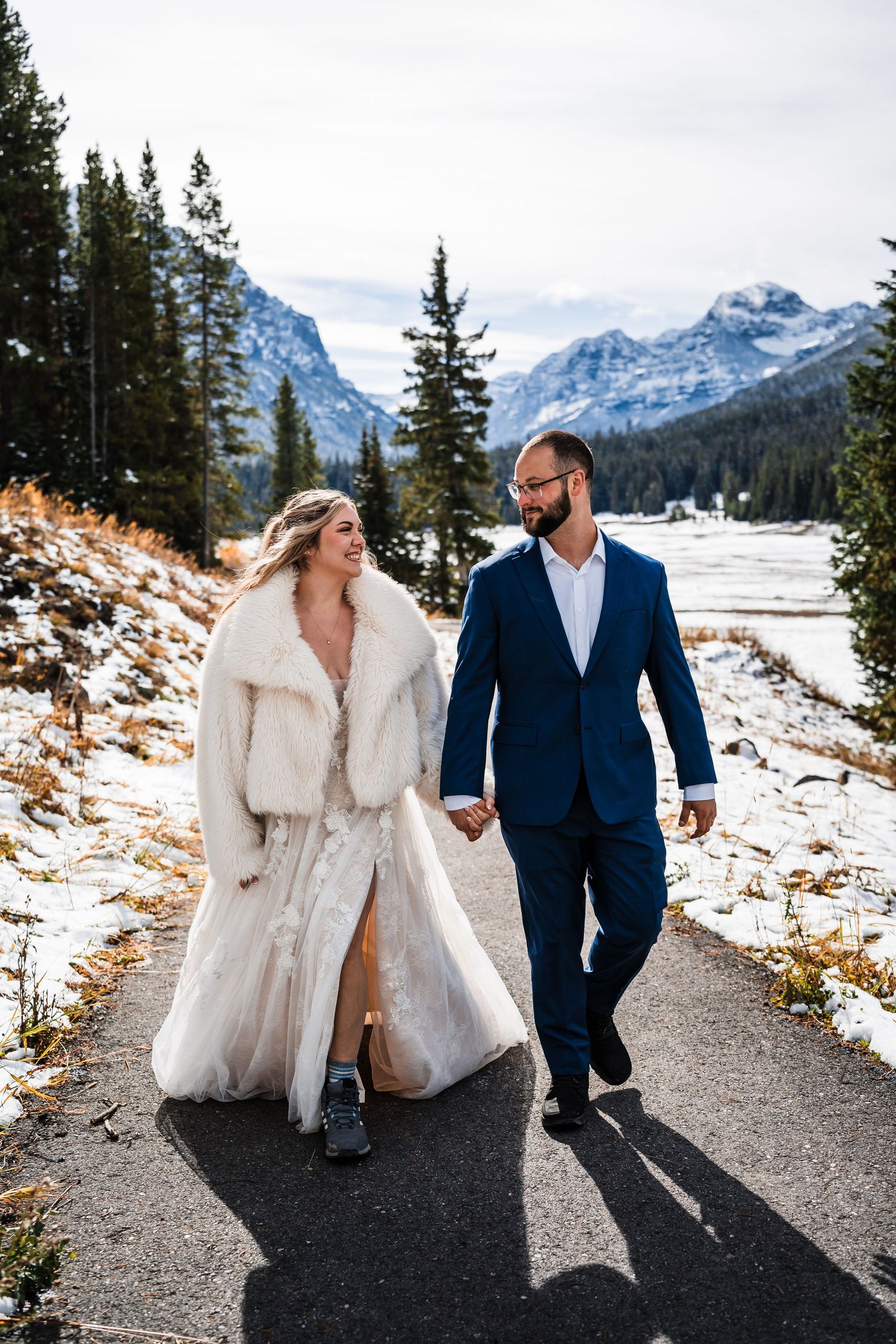 A couple walking on a snowy mountain path with evergreen trees and snow-capped peaks in the background.