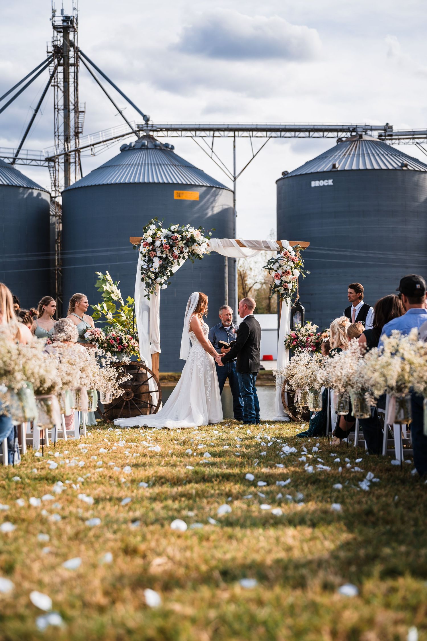 Outdoor wedding ceremony setup with grain silos in background and floral decorations.