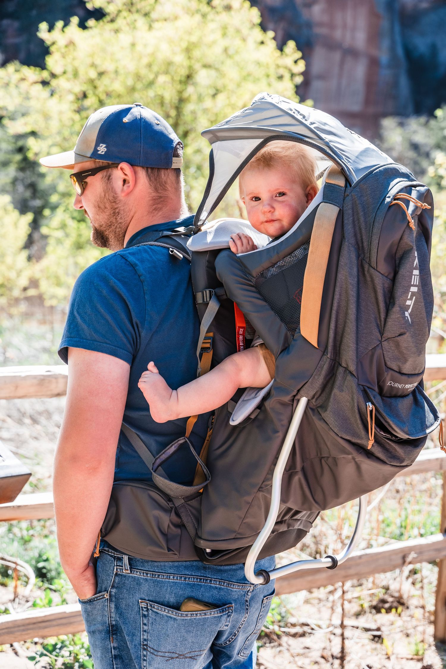 Adult wearing a blue shirt carries a small child in a gray hiking backpack carrier on an outdoor trail.