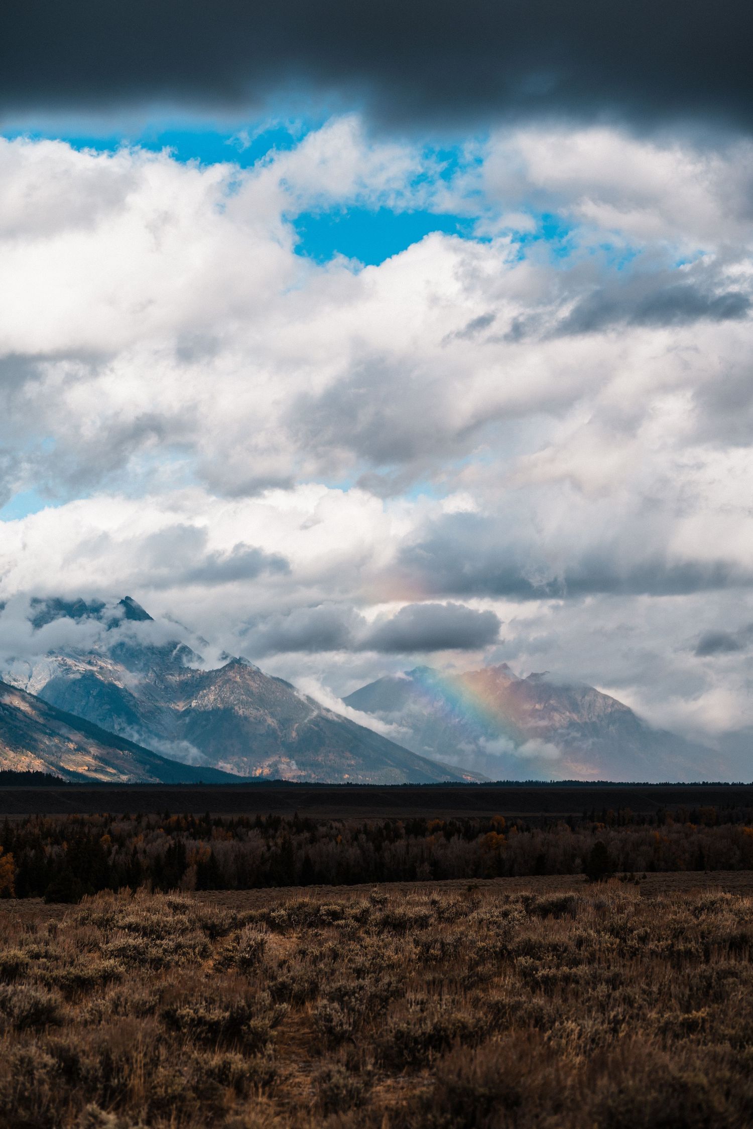 Dramatic mountain landscape with snow-capped peaks under moody clouds stretching across a vast brown prairie field.