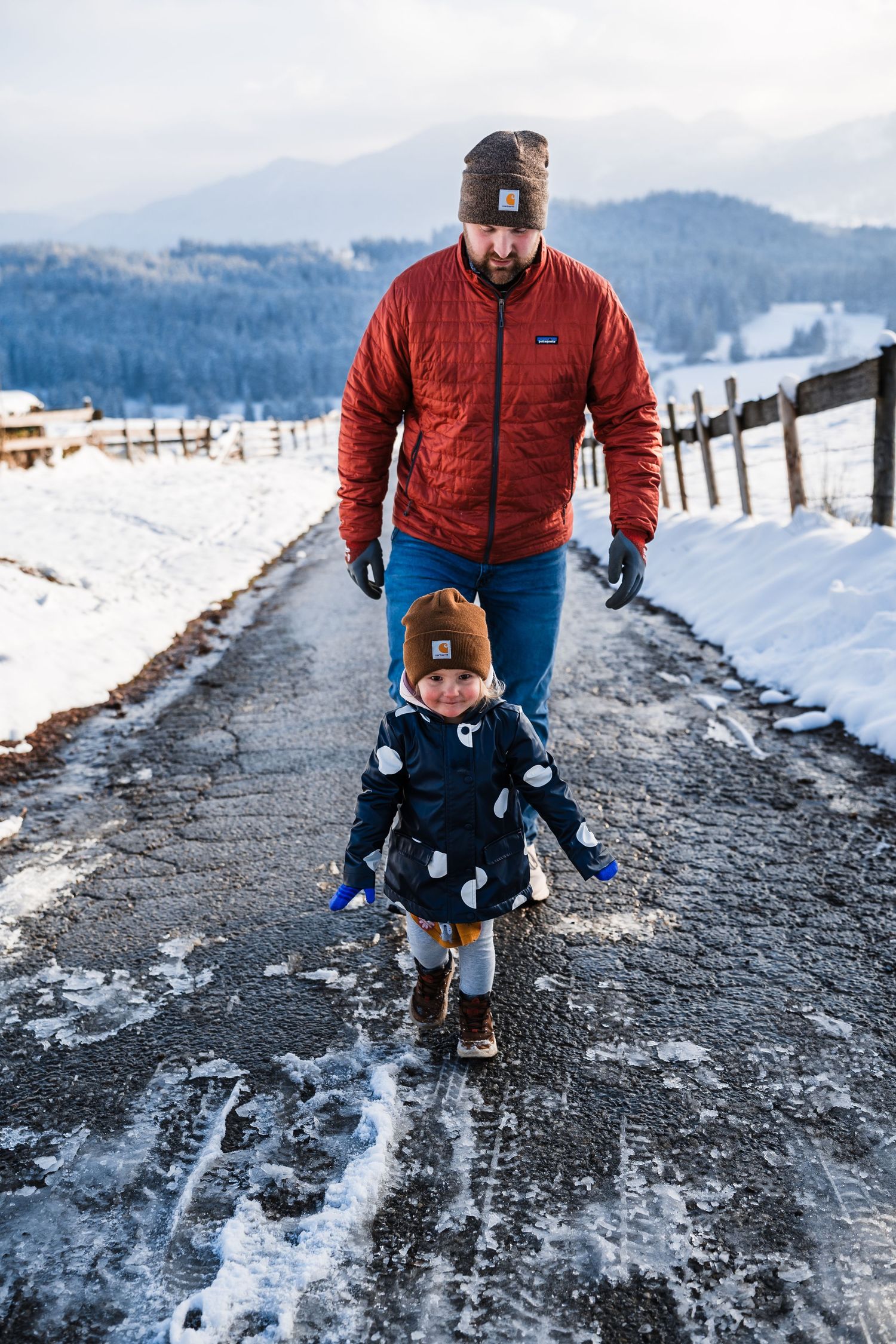 Two people walk along a snowy mountain path with scenic winter views in the background.
