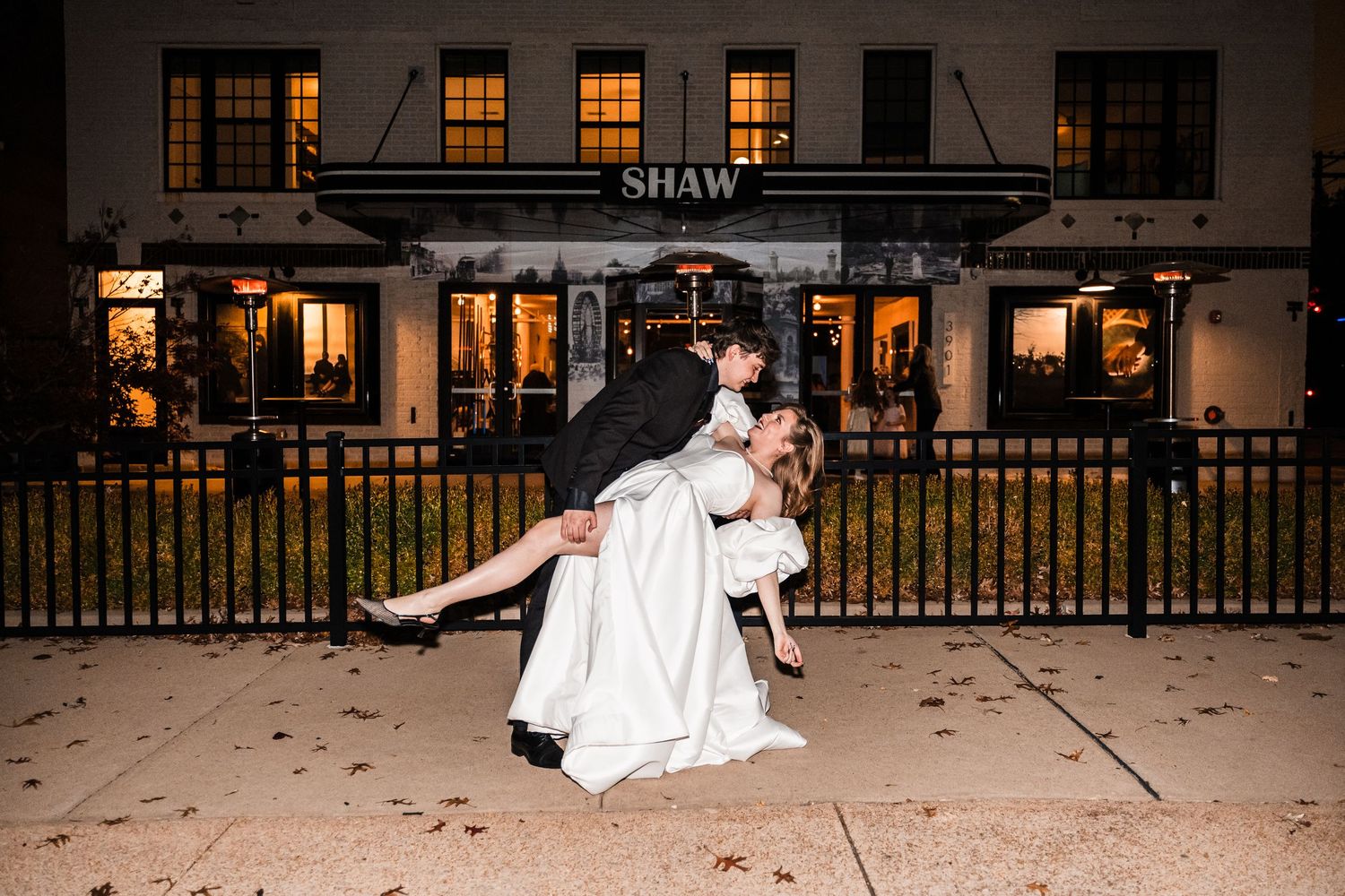 A wedding couple dances together at night on a patio with glowing windows and scattered leaves in the background.