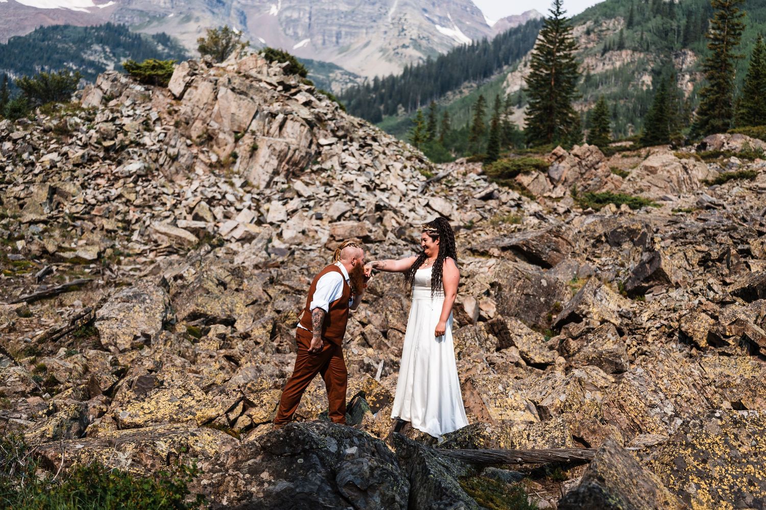 A groom helps his bride navigate rocky terrain with mountain peaks and pine trees in the background.