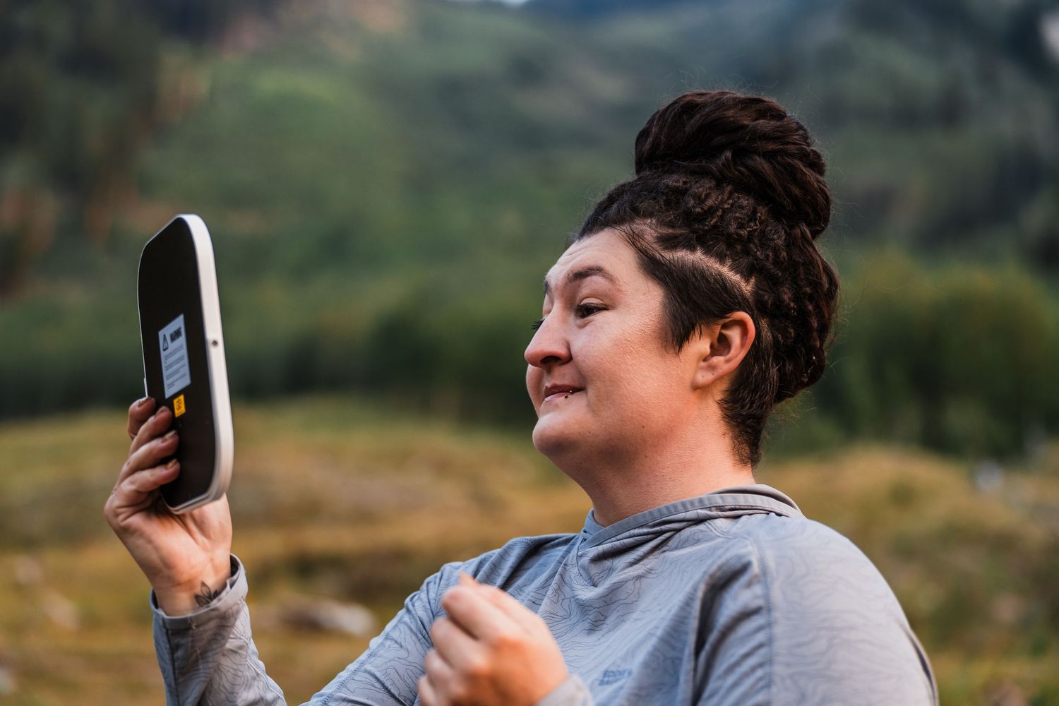 Woman smiling while holding a mobile WiFi hotspot device outdoors with green hills in the background.