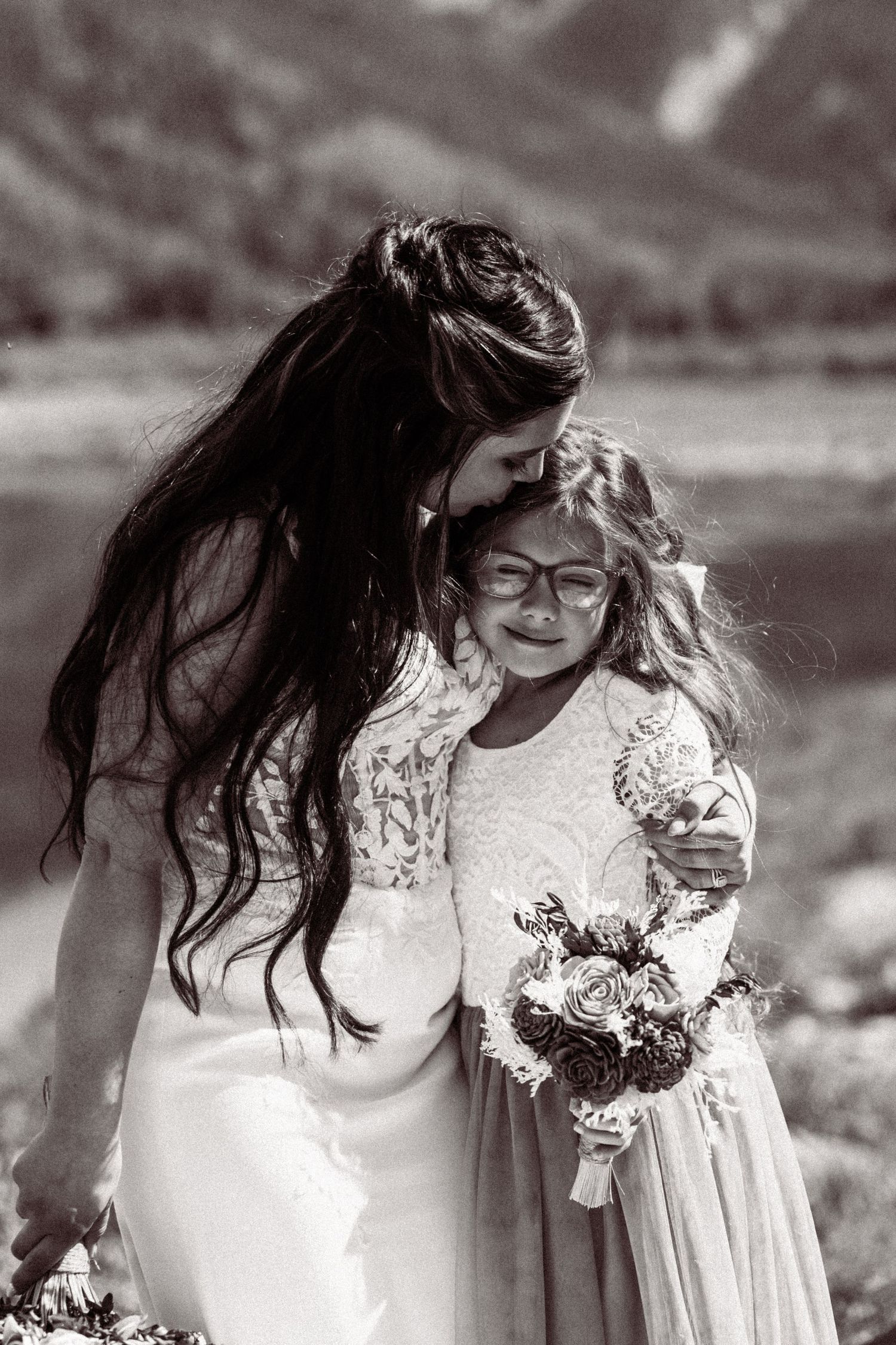 Bride in lace gown embraces emotional flower girl holding bouquet in black and white outdoor portrait.