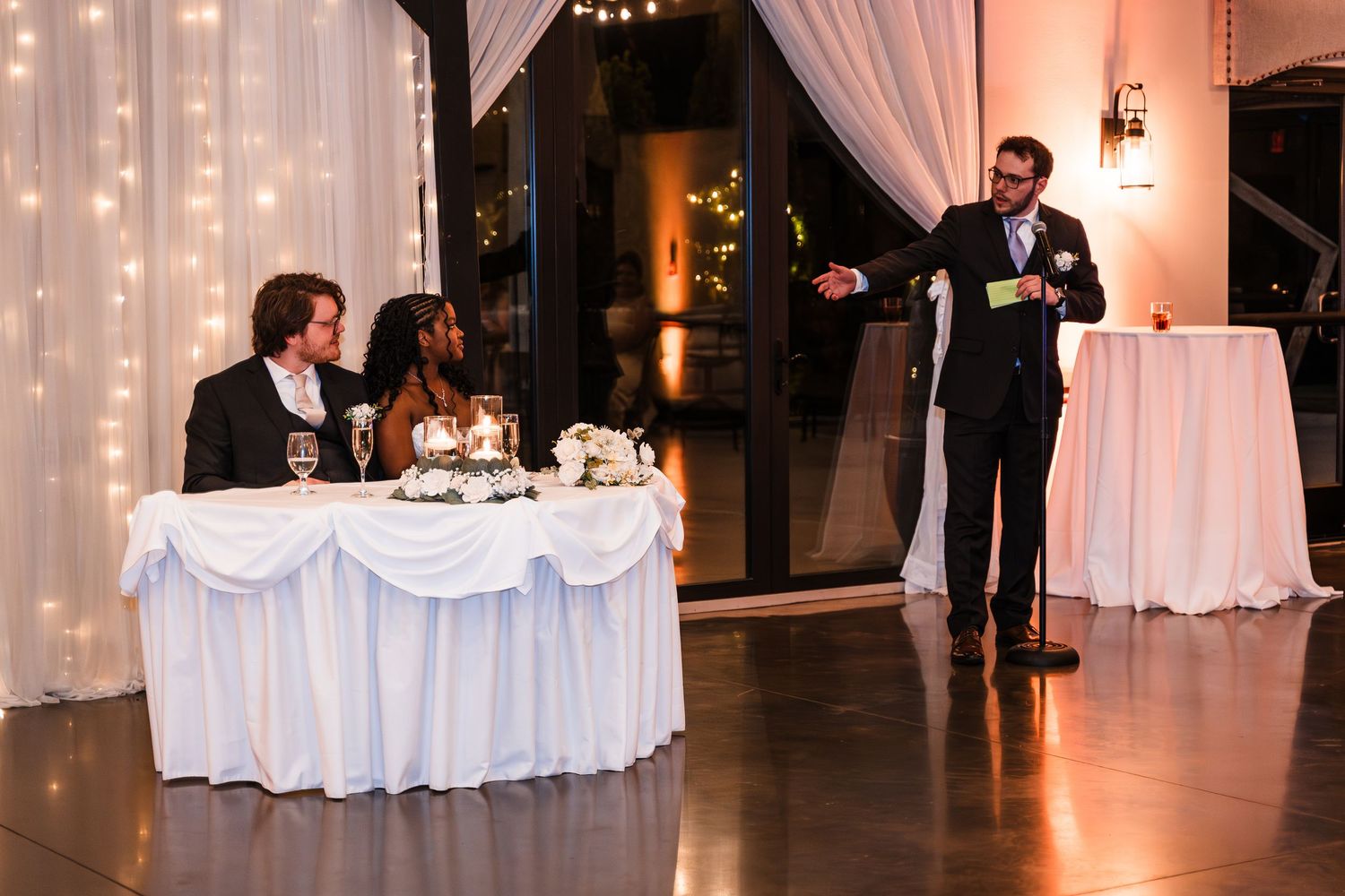 Best man delivers toast to seated bride and groom at elegantly decorated wedding reception.