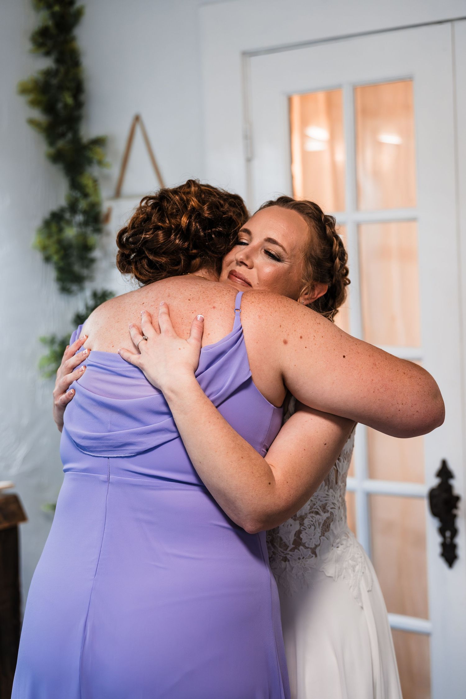 Bride in white lace gown shares emotional hug with bridesmaid in lavender dress during wedding day preparations.