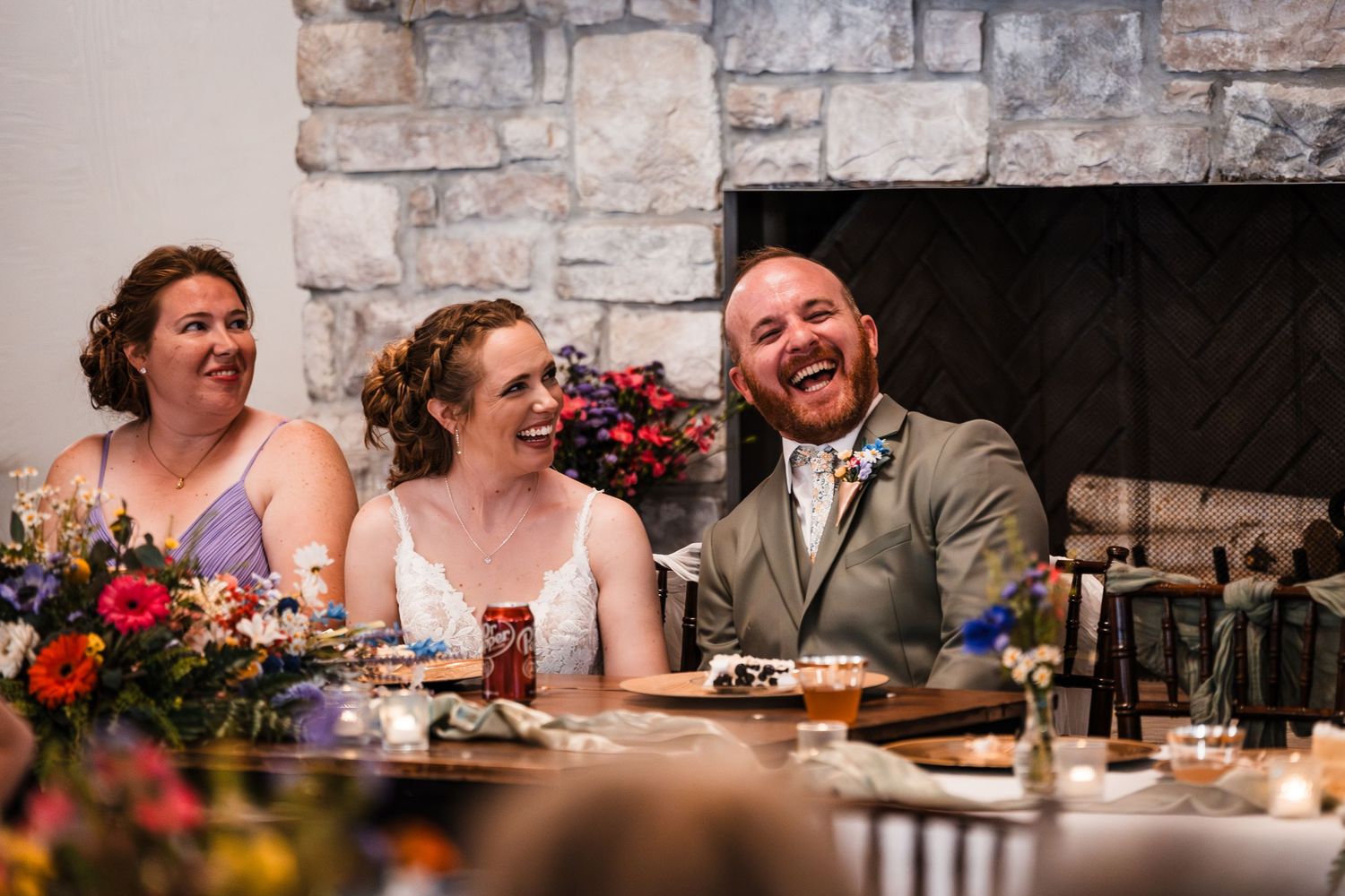 Bride and groom laugh joyfully at their wedding reception near a stone fireplace.