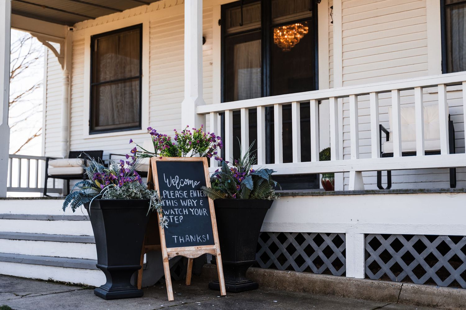 White Victorian farmhouse porch entrance with chalkboard welcome sign and black planters filled with purple flowers.