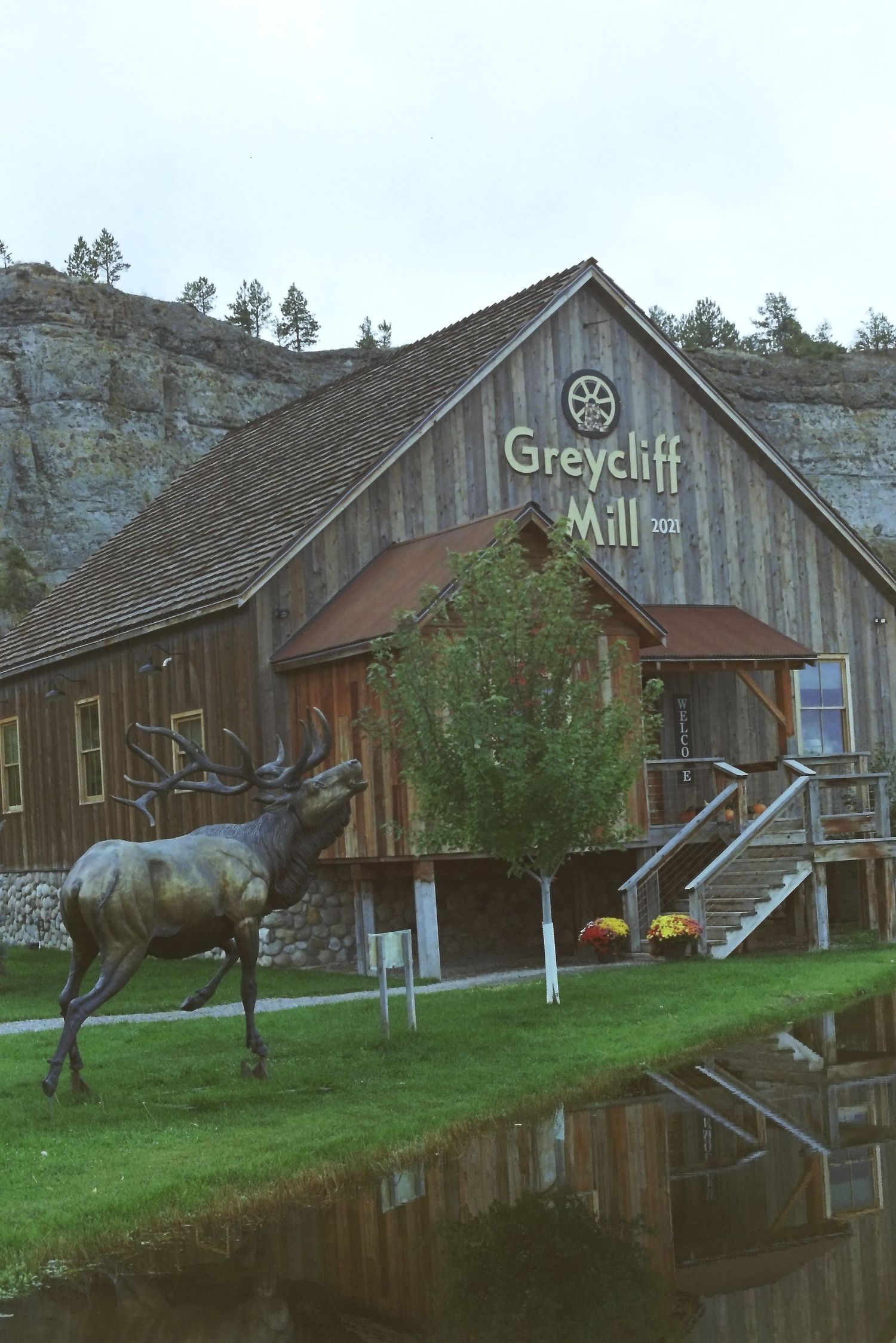 Historic Greycliff Mill buildings stand along a tranquil pond with rustic wooden structures reflecting in the calm water.