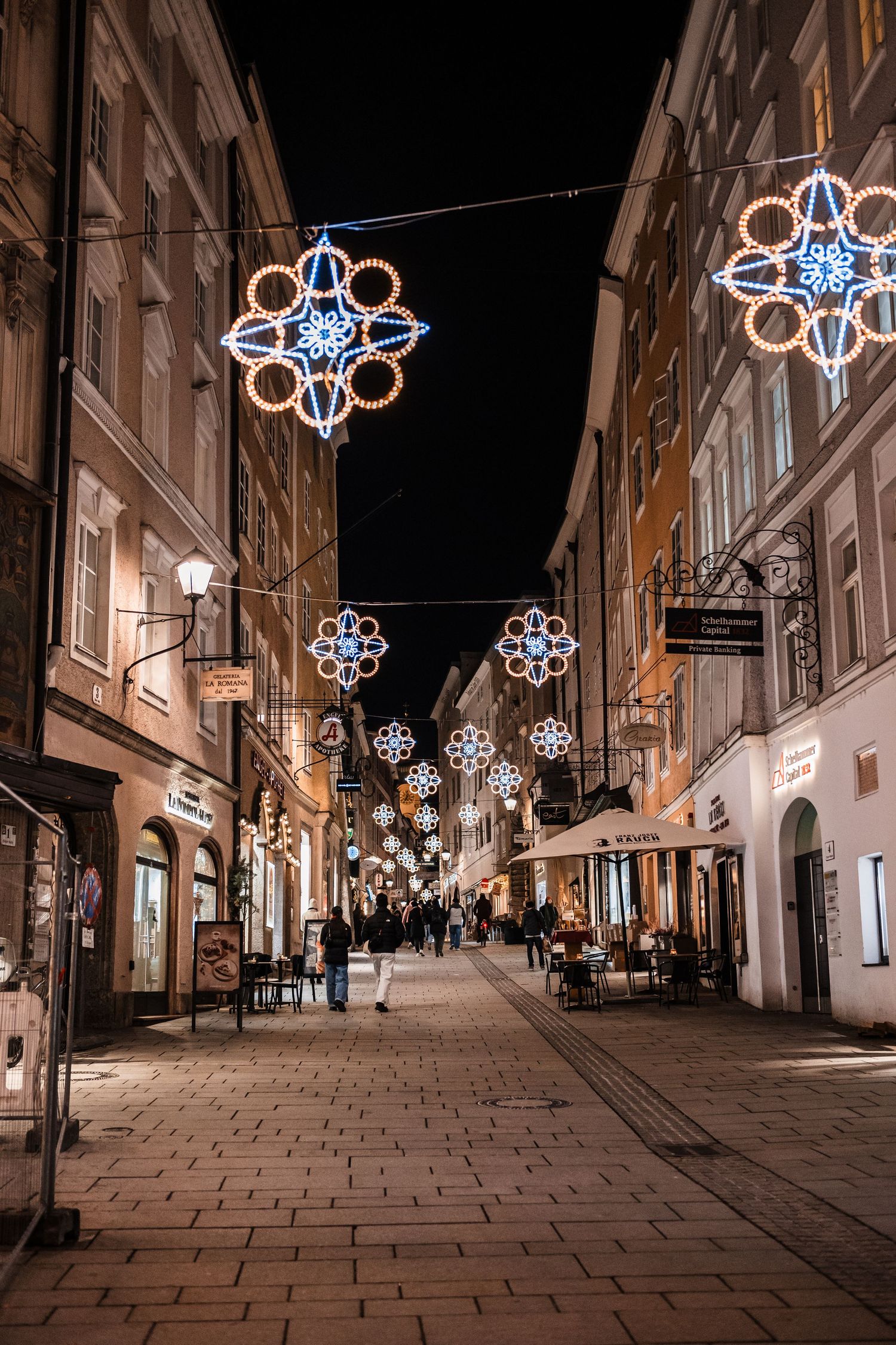 Illuminated Christmas decorations hang over a cobblestone street in a historic European city at night.