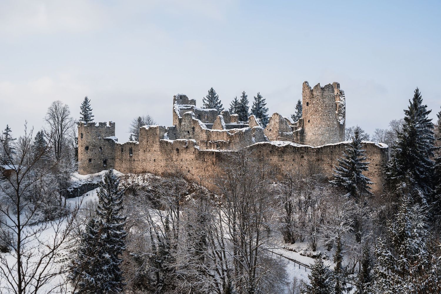 Ancient snow-covered castle ruins perched on a hilltop surrounded by evergreen trees in winter.