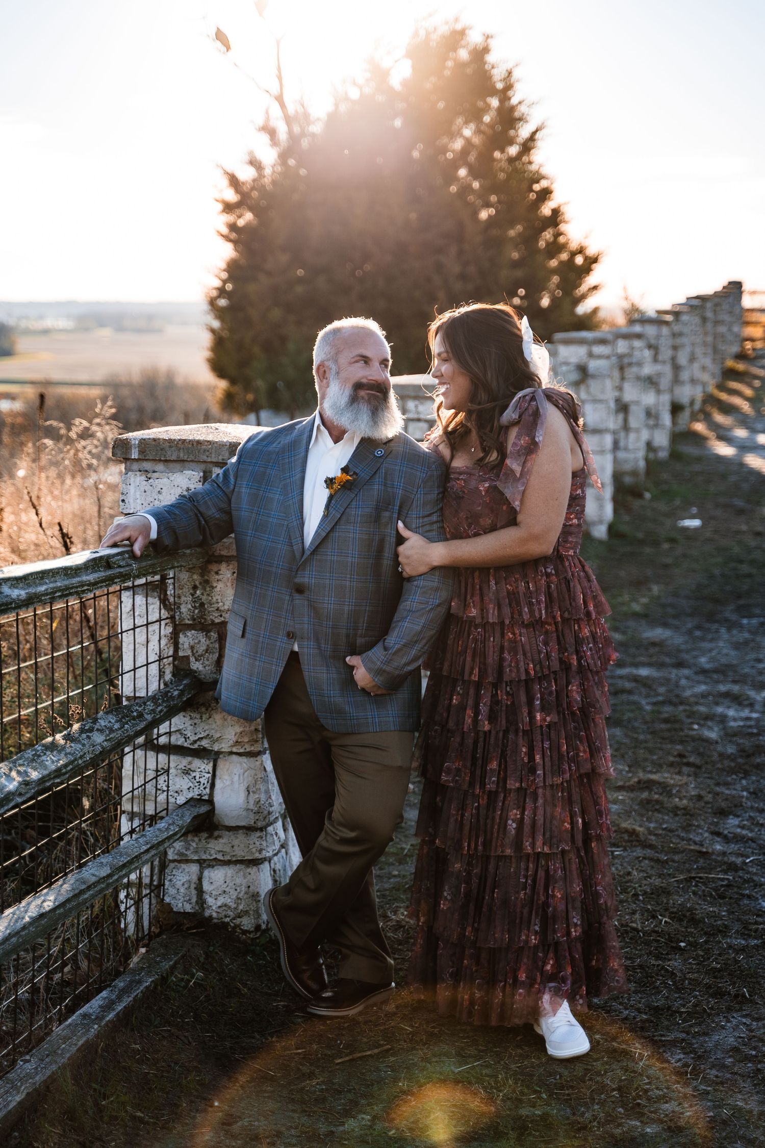 Couple in formal wear share intimate moment by stone wall during sunset.