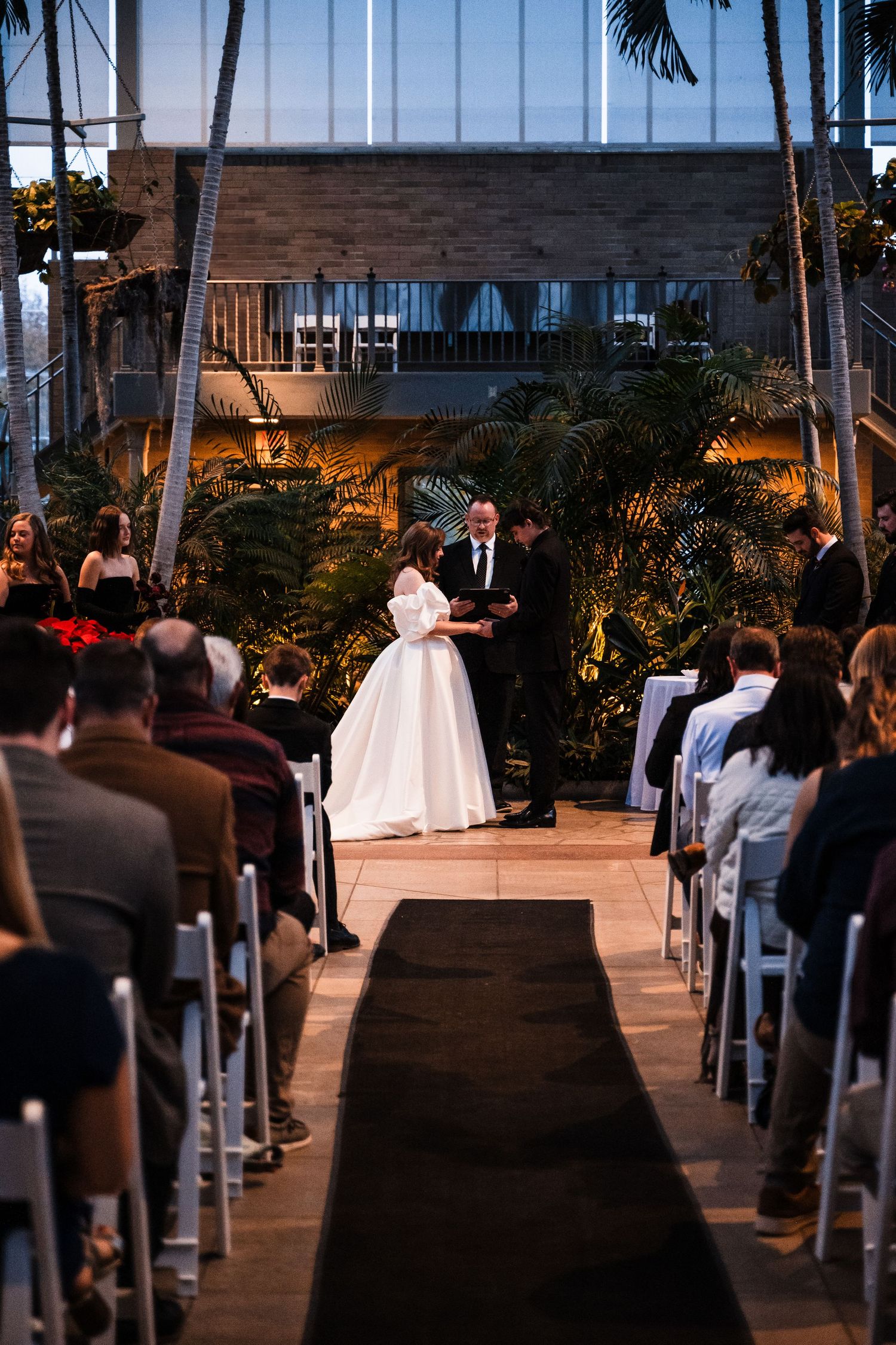 Evening wedding ceremony in a palm-filled venue with guests seated along a black-carpeted aisle.