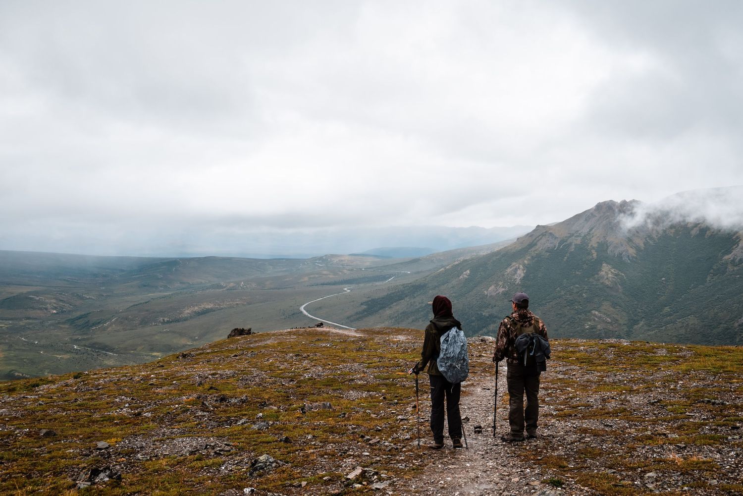 Two hikers observe the mountainous landscape from a hiking trail summit.
