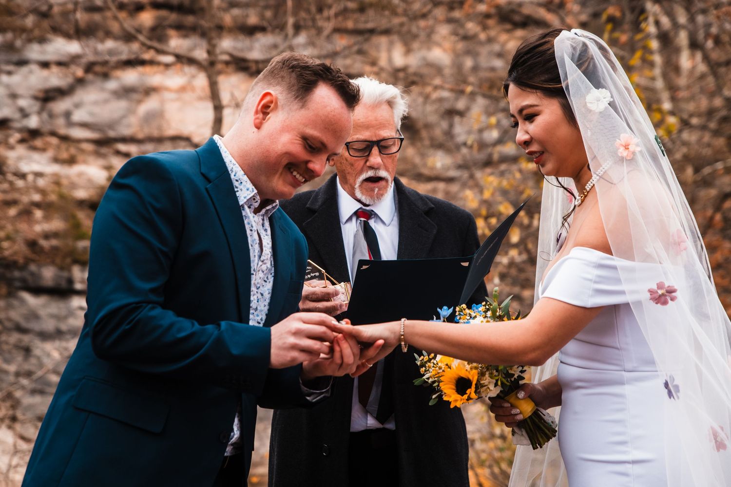 Emotional exchange of rings during intimate outdoor wedding ceremony with officiant present.