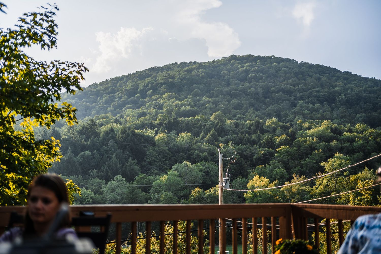 A scenic mountain vista viewed through a wooden deck railing with trees and a church steeple in the distance.