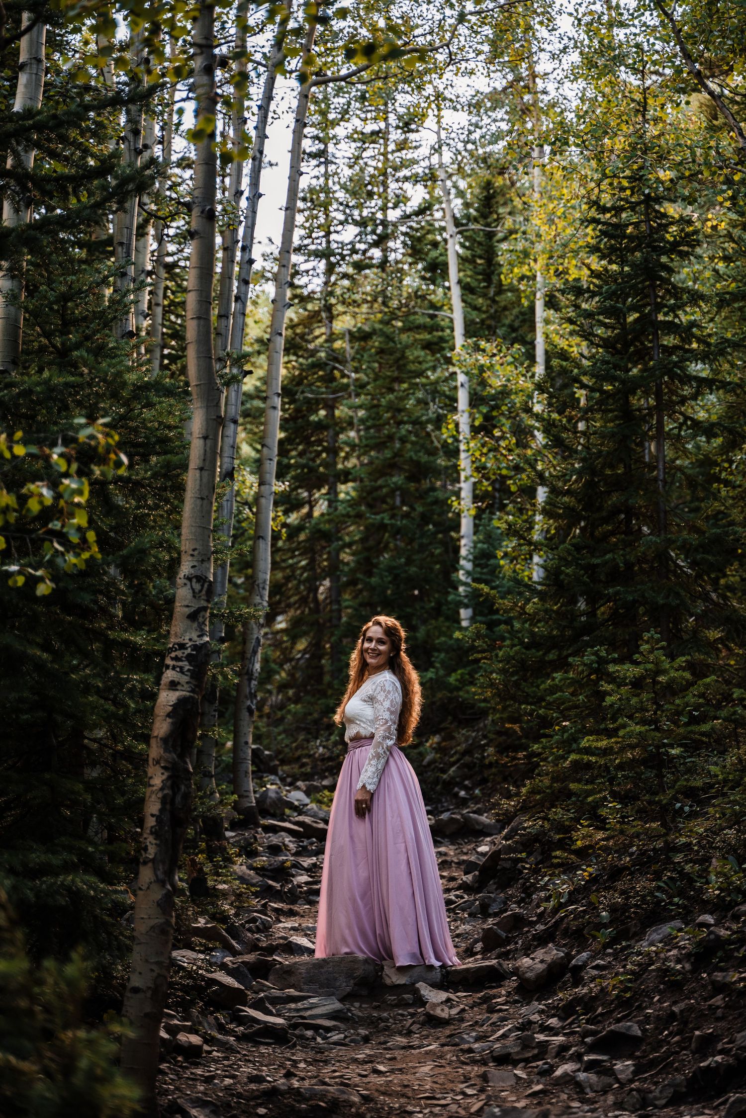 Pink-dressed figure stands on forest trail surrounded by tall aspen trees.