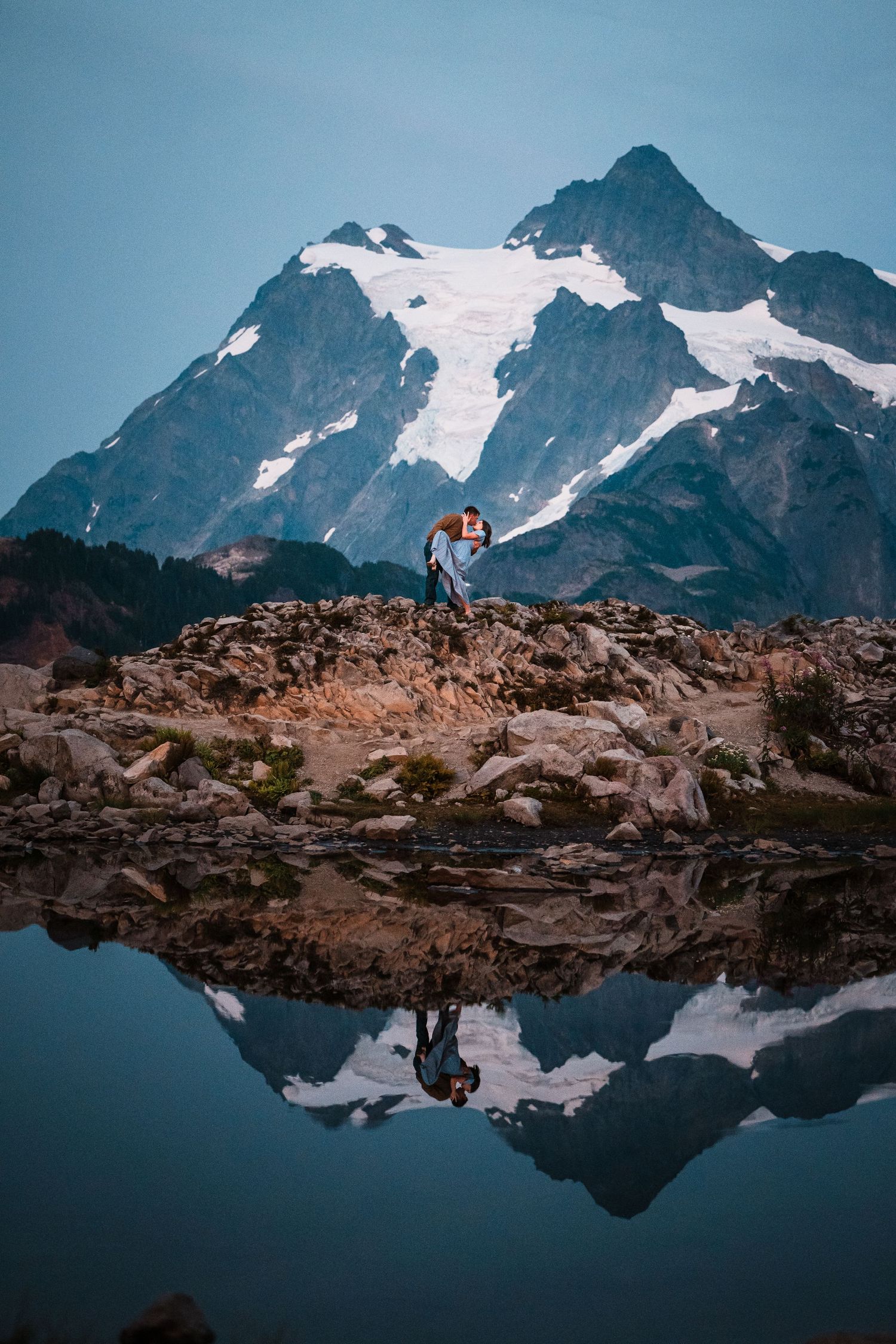 Snow-capped mountain peaks reflect in a calm alpine lake with two small figures visible on the rocky shore.