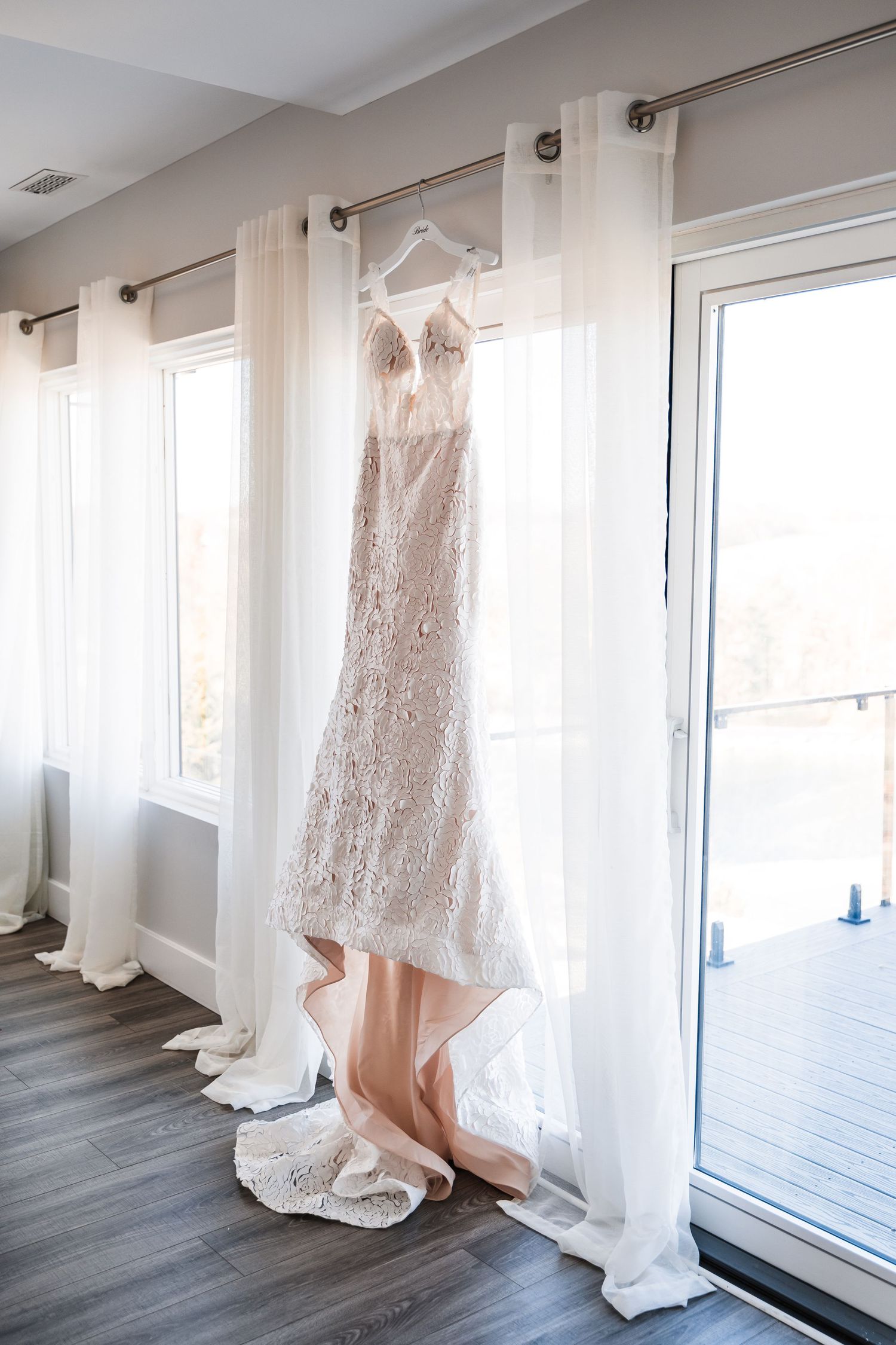 A white lace wedding dress hangs elegantly by a bright window with natural light streaming in.