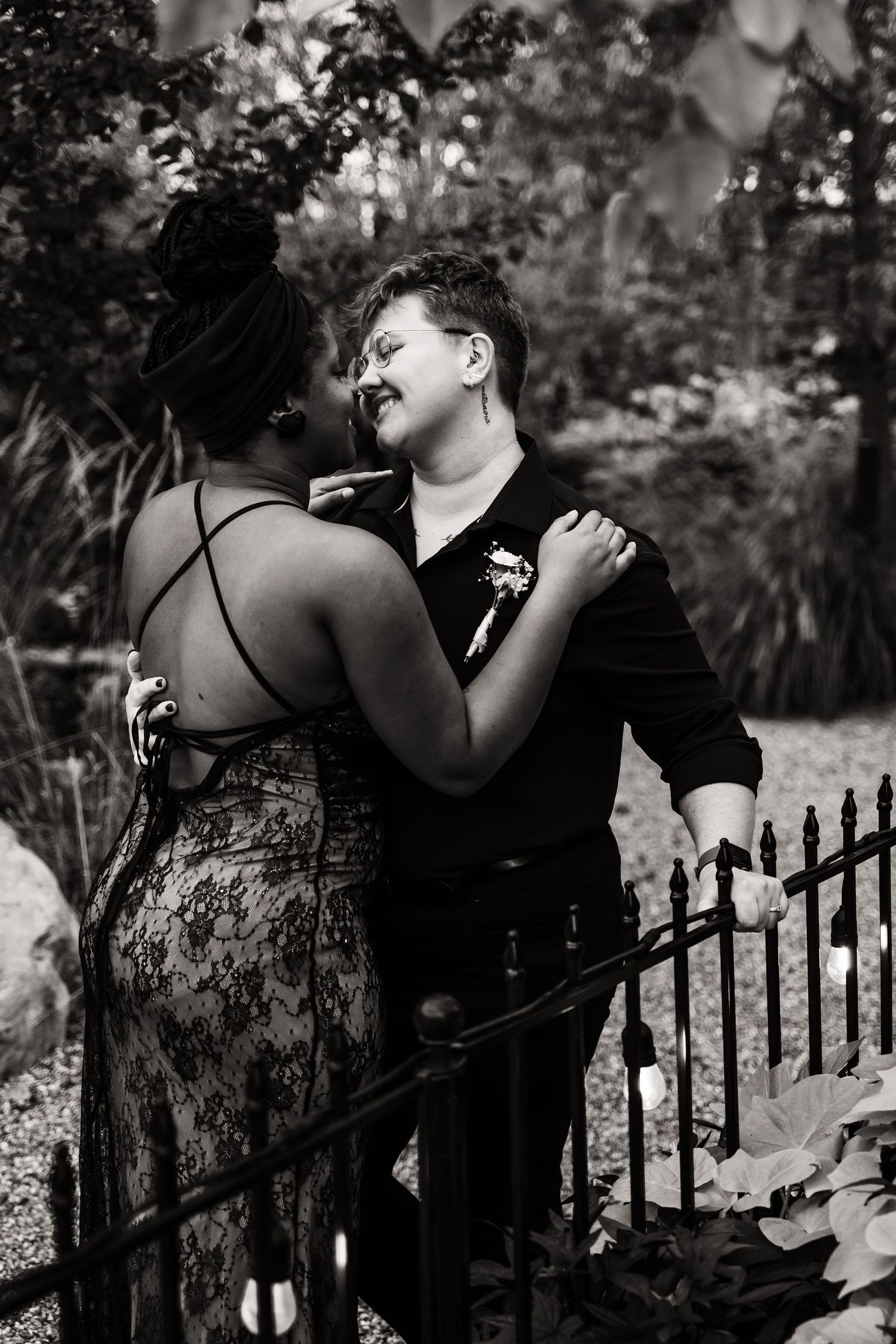 A romantic black and white portrait of a couple embracing by an iron fence in a garden setting.