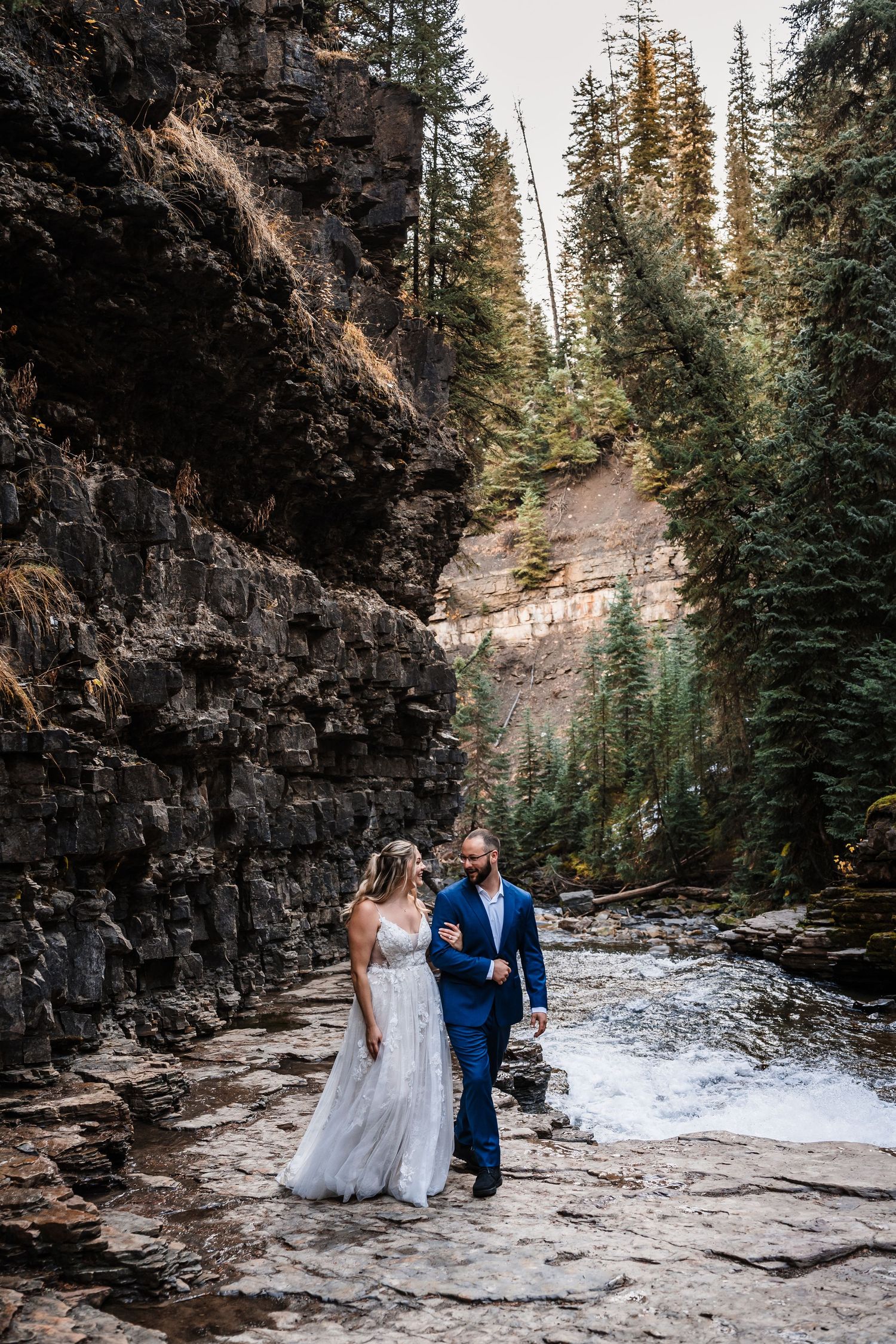 A wedding couple poses together on rocky terrain surrounded by evergreen trees and cliffs.