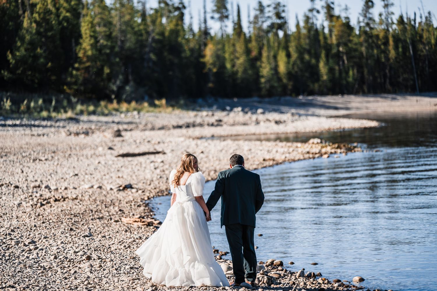 A couple walks along a scenic lakeside beach surrounded by evergreen forest on a sunny day.