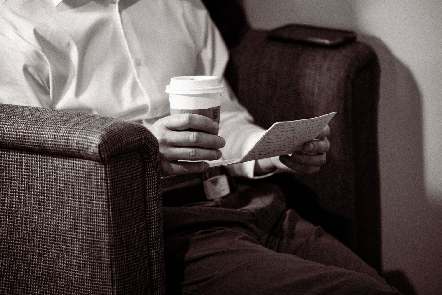 Man in white shirt sitting in armchair holding coffee cup and phone in black and white photo.