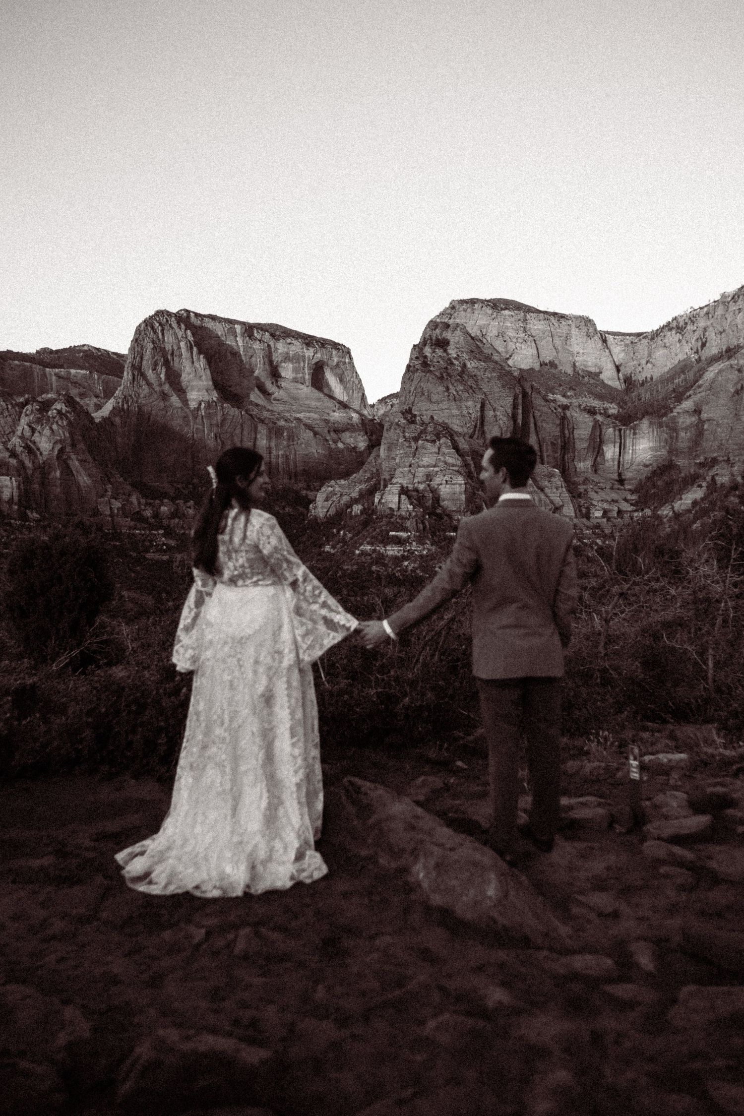Bride and groom holding hands gazing at dramatic desert rock formations at dusk.