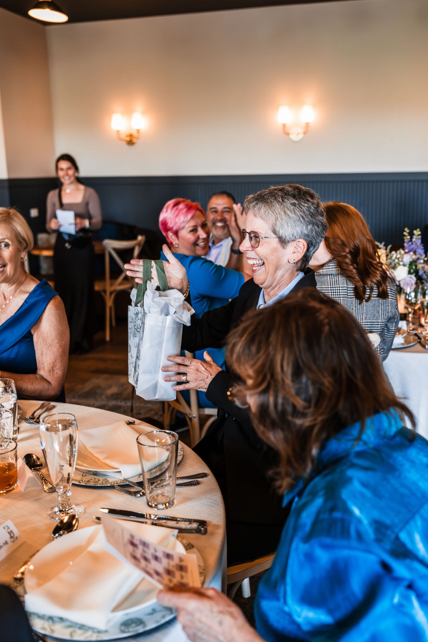 Guests laughing and exchanging gifts at an elegant indoor wedding reception dinner party.