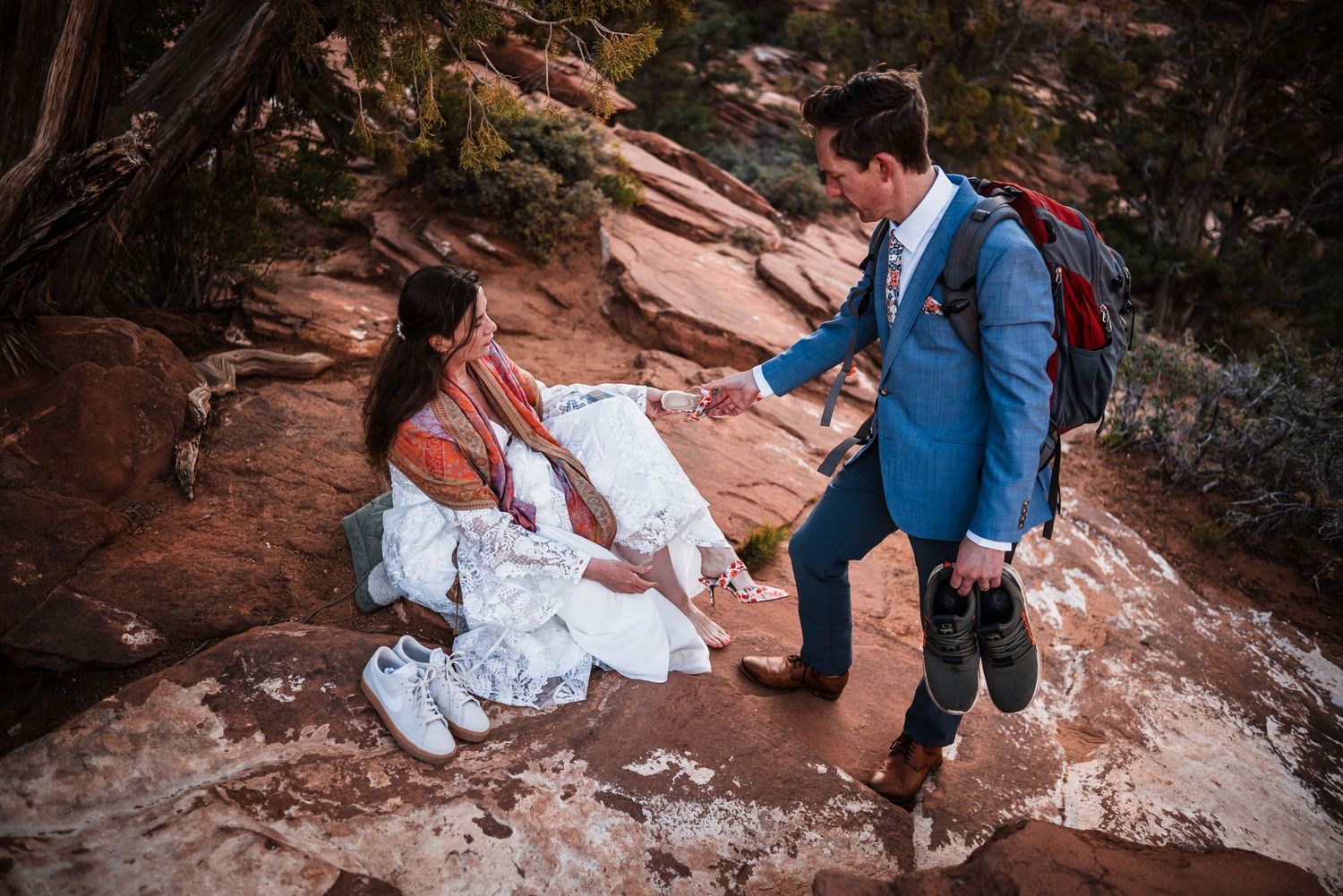 Groom in blue suit helps bride in lace wedding dress navigate red rock terrain during outdoor elopement.