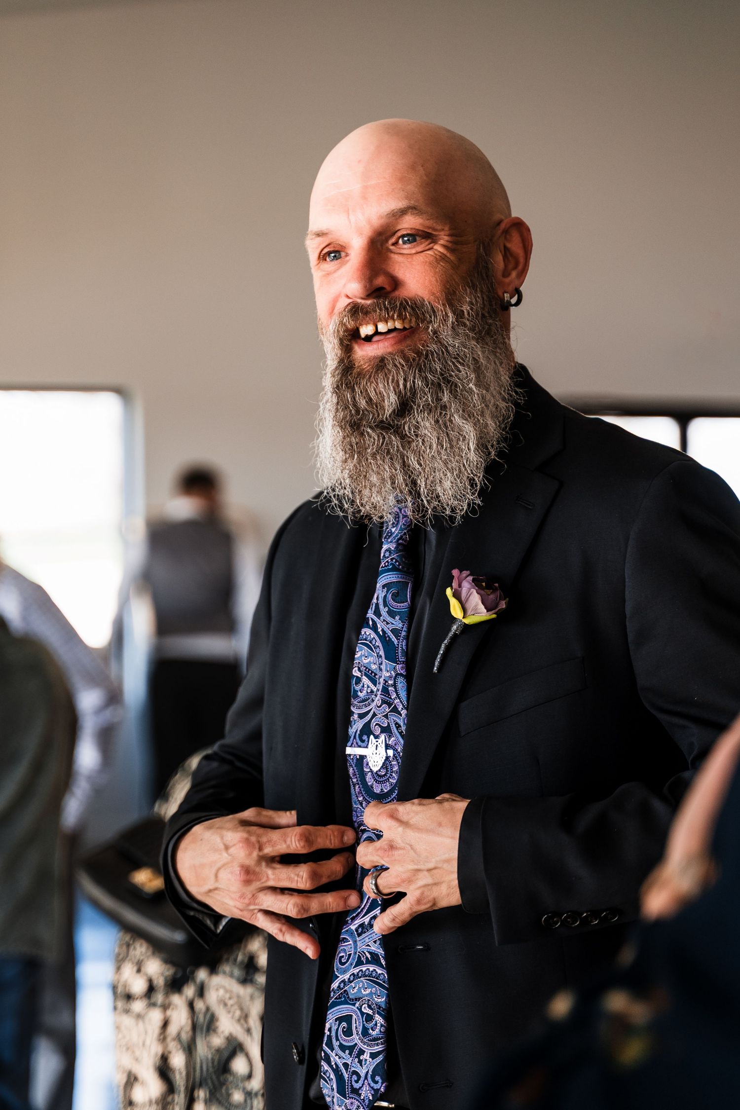 Smiling bald groom with long beard wears black suit and paisley tie with boutonniere.