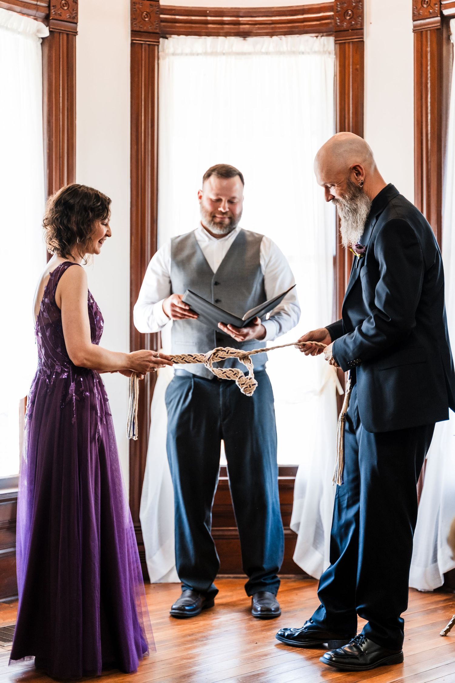 Couple performs rope tying unity ceremony with officiant during intimate indoor wedding.