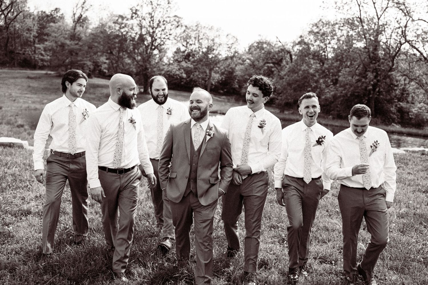 Black and white photo of groom and groomsmen in gray suits walking together across a grassy field.