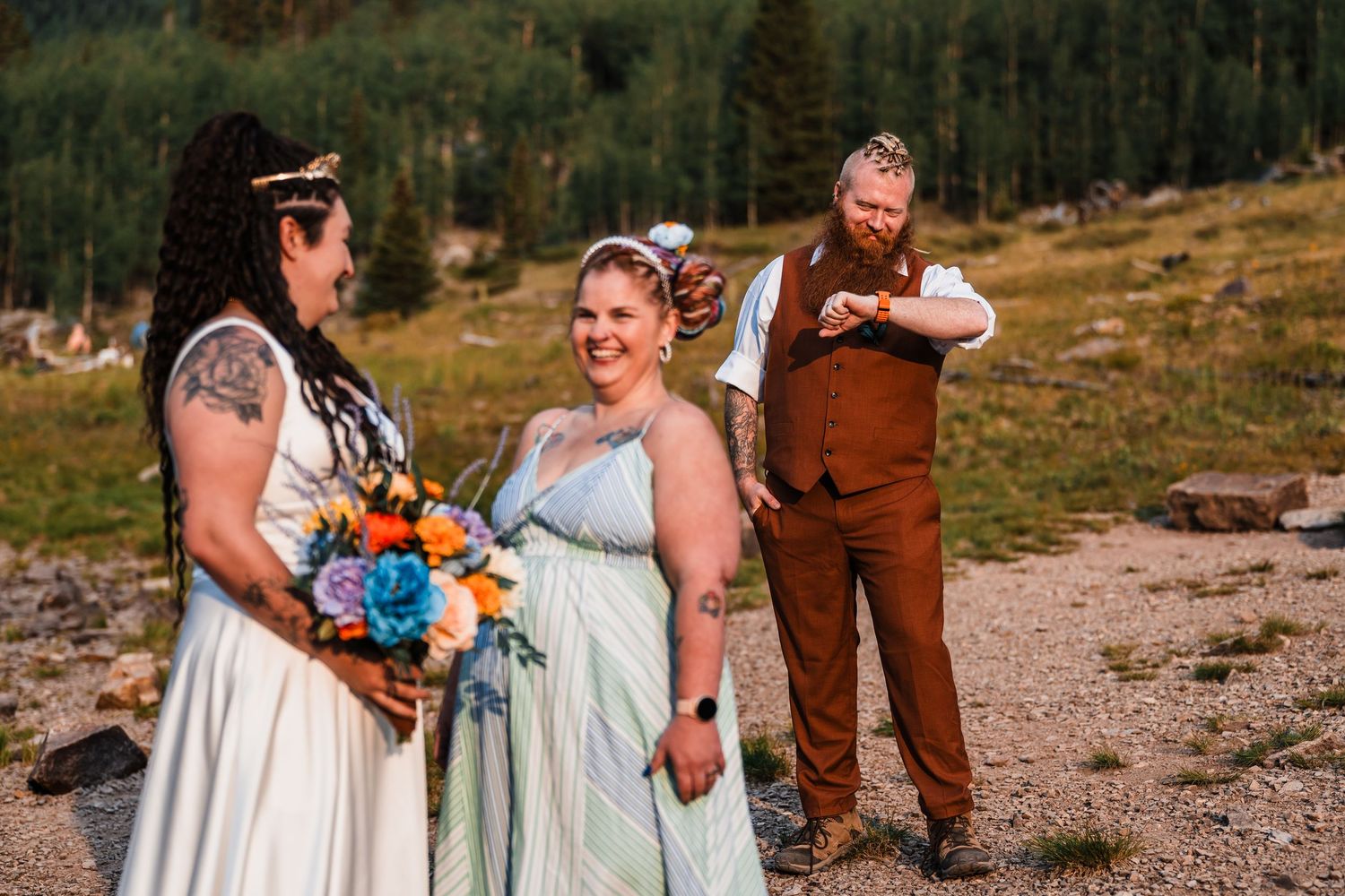 Two brides hold colorful bouquet while officiant reads vows at outdoor mountain elopement ceremony.