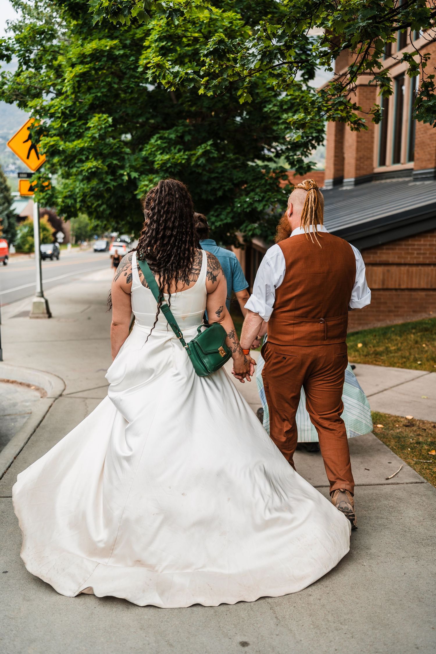 Bride in white gown with green bag and groom in brown vest walk hand in hand down a tree-lined sidewalk.
