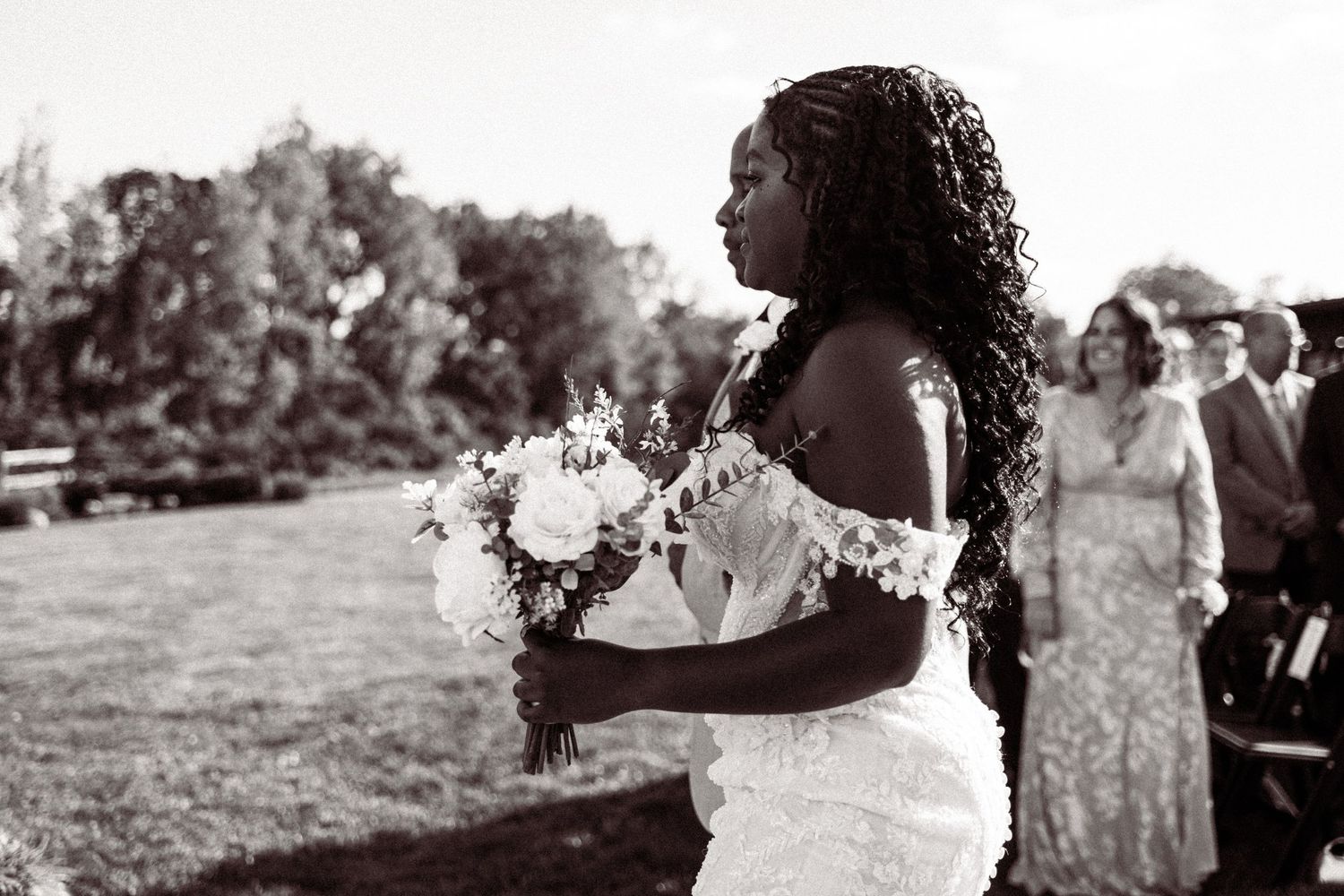 A Black bride with braided hair holds a white floral bouquet at an outdoor wedding ceremony in black and white.