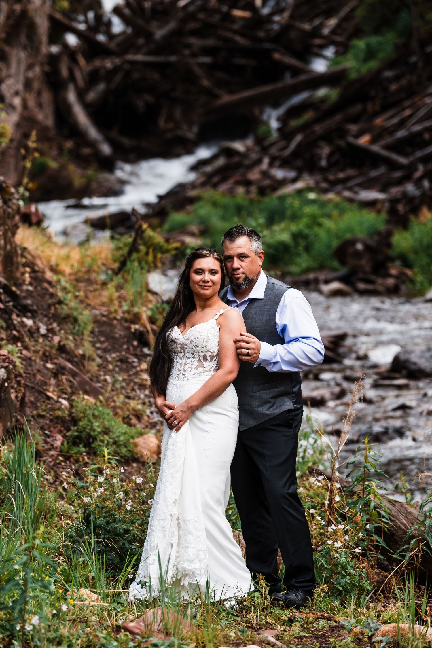 Bride in lace wedding gown and groom in gray vest embracing near a rocky mountain stream.