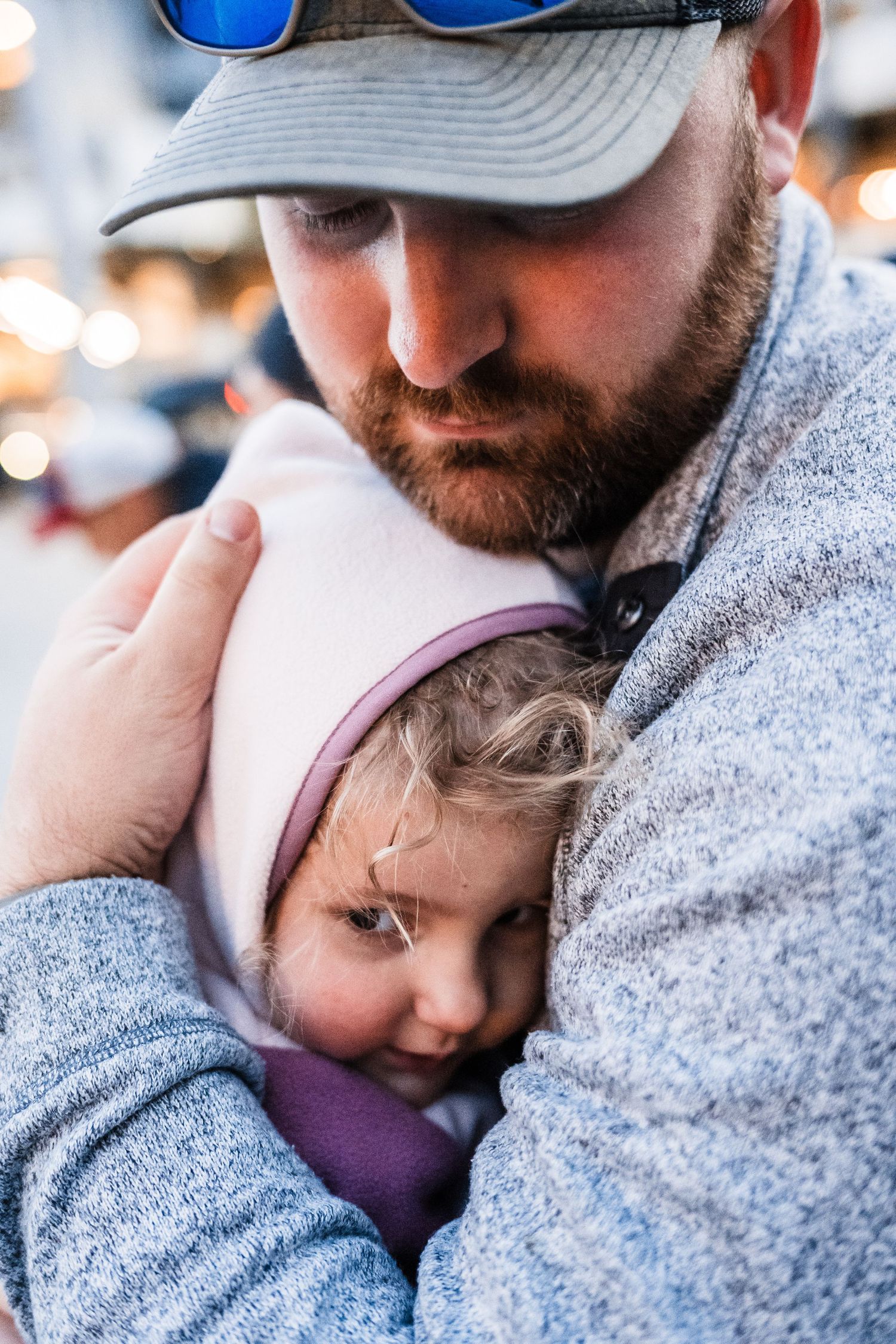 A tender moment shows a child resting peacefully against a person wearing a gray sweater and baseball cap.