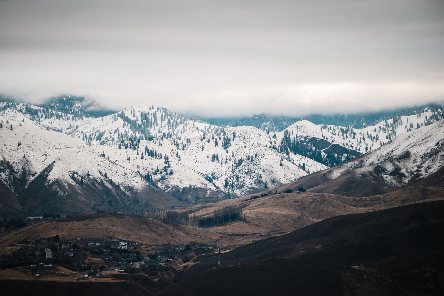 Snow-capped mountain range towers over brown valley with small buildings visible below.