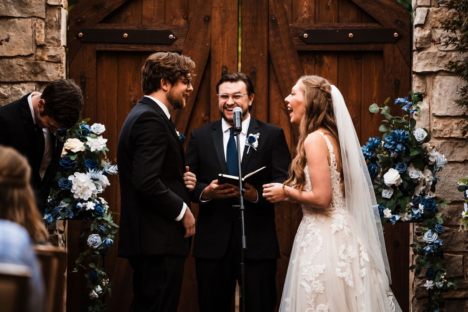 Wedding ceremony in front of rustic wooden doors decorated with blue and white flowers.
