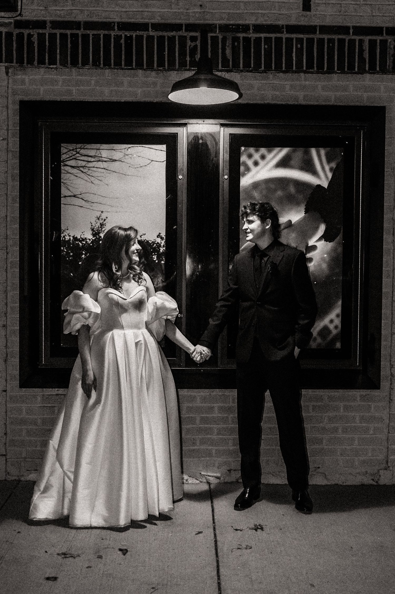 Black and white photo of a couple in formal attire standing outside under a vintage storefront light.
