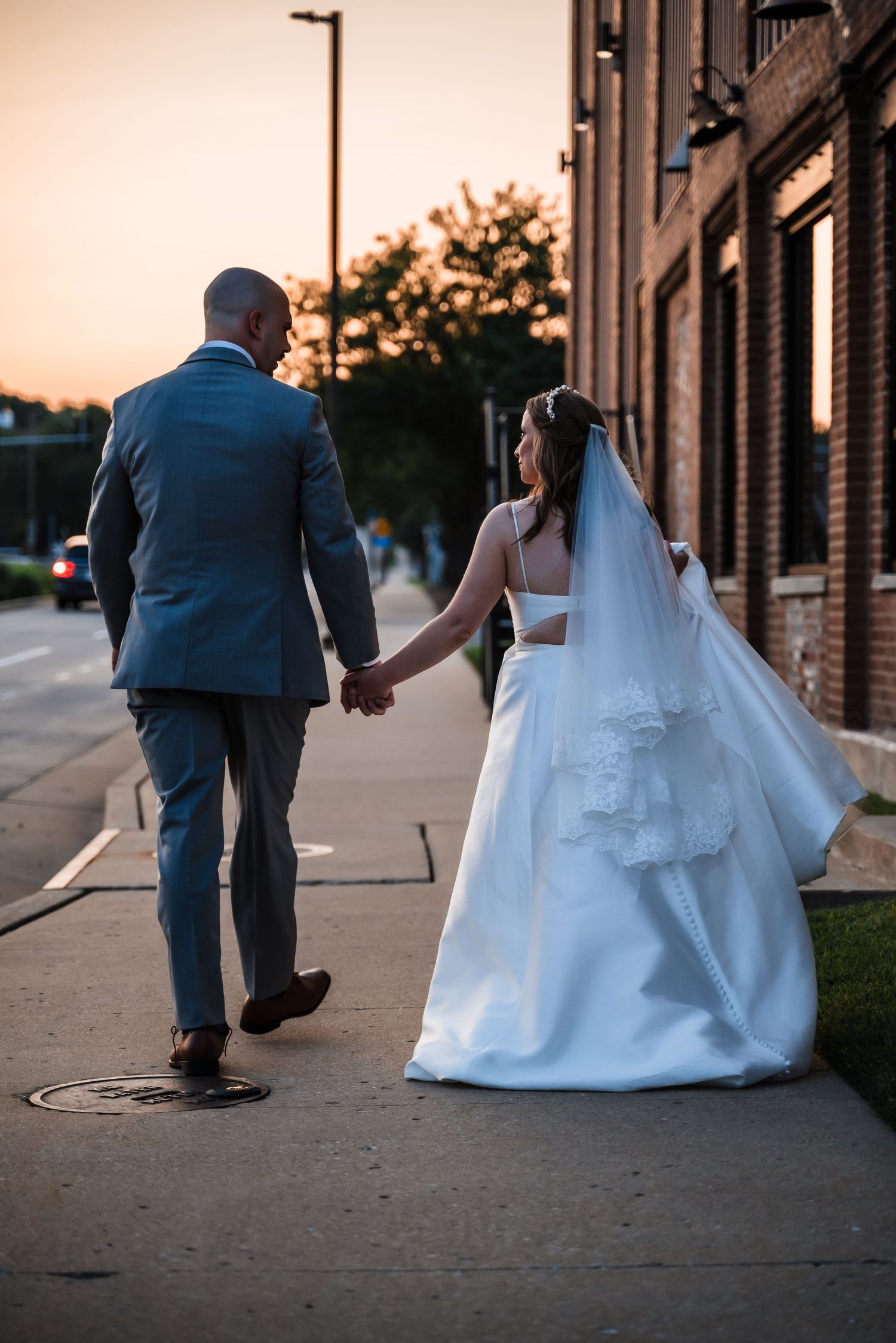 Newlyweds walking hand in hand down a city sidewalk at sunset.