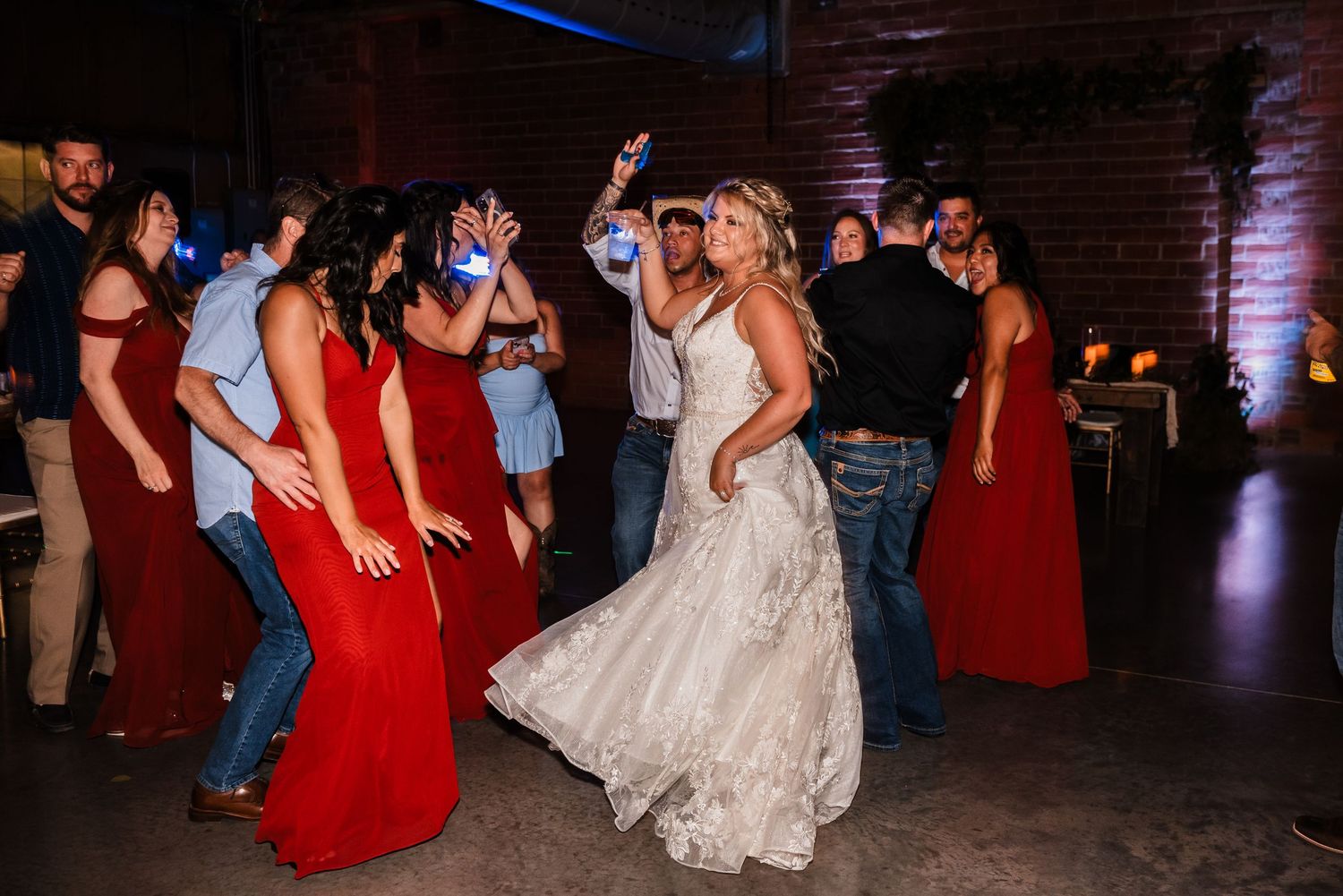 Wedding party guests in red dresses and jeans dance with a bride in a white lace gown at a dimly lit indoor reception venue.