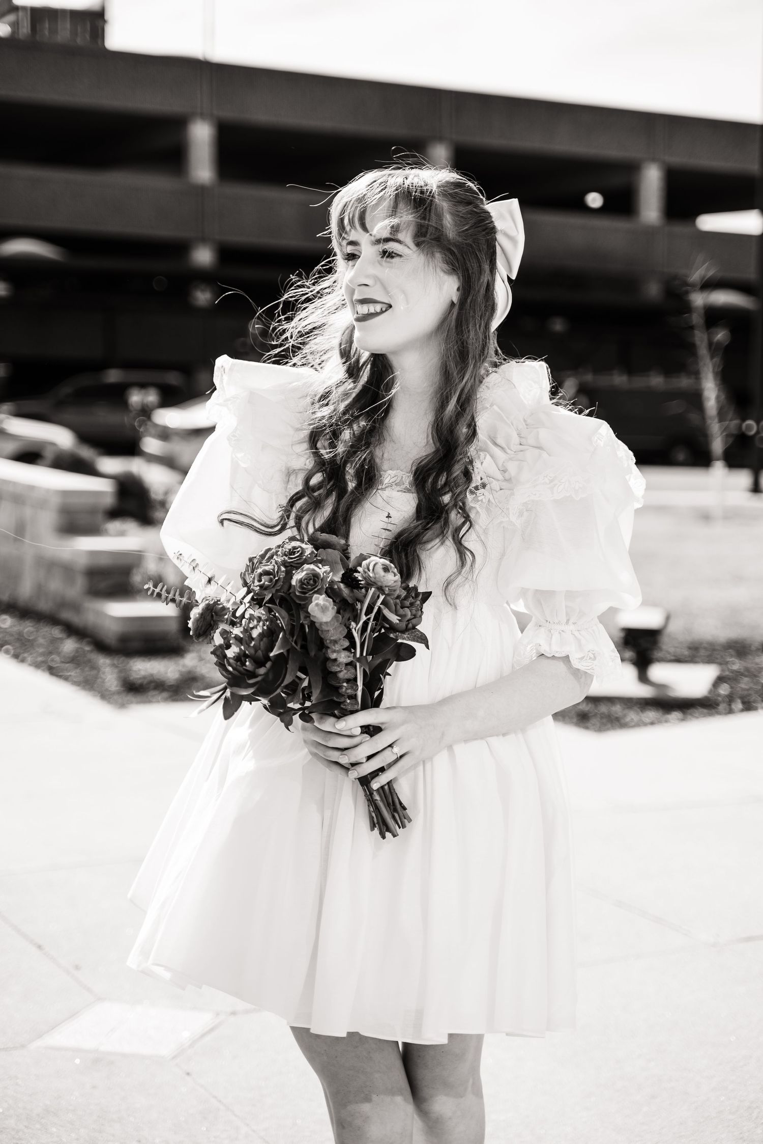 A bride in a short white dress holds a bouquet while standing outdoors.