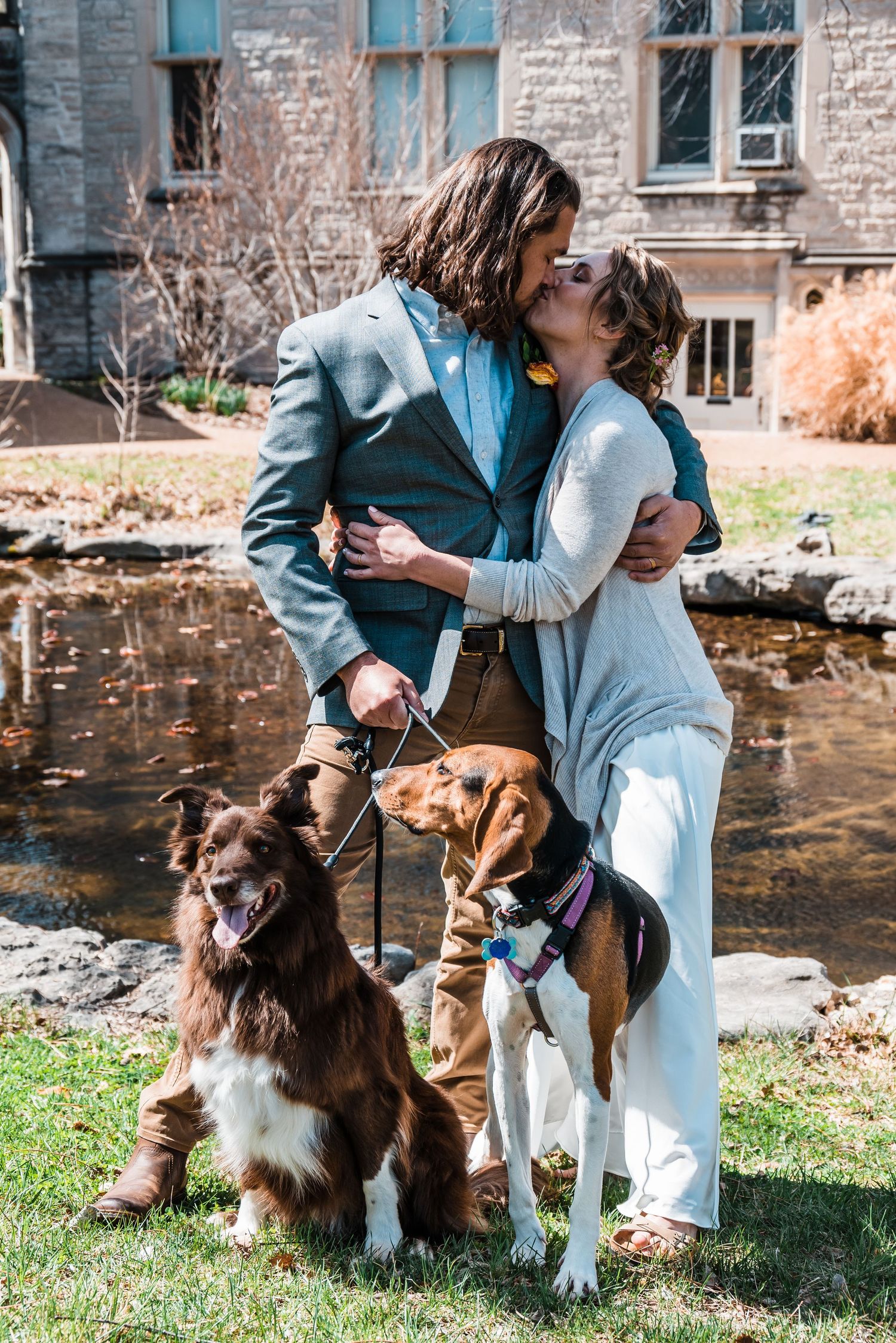 Romantic couple with two dogs sharing tender moment by stone building and pond.